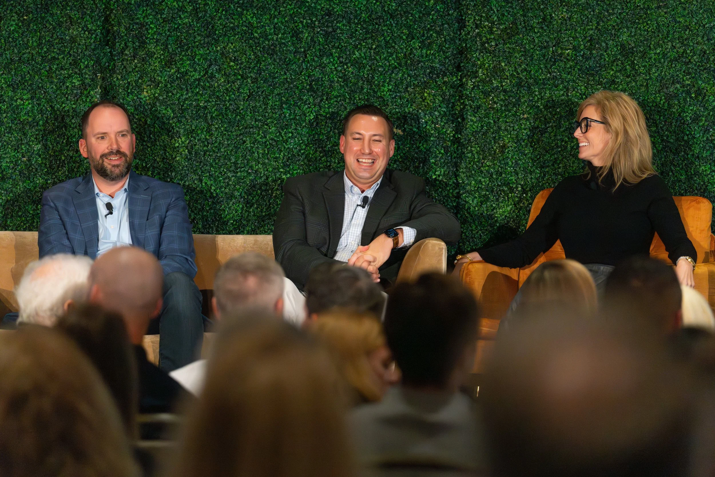 Three people sitting on a stage in front of an audience, engaging in a panel discussion. The woman on the right is smiling and wearing glasses, while the man in the middle is laughing, shaking hands with the woman. The man on the left has a beard and