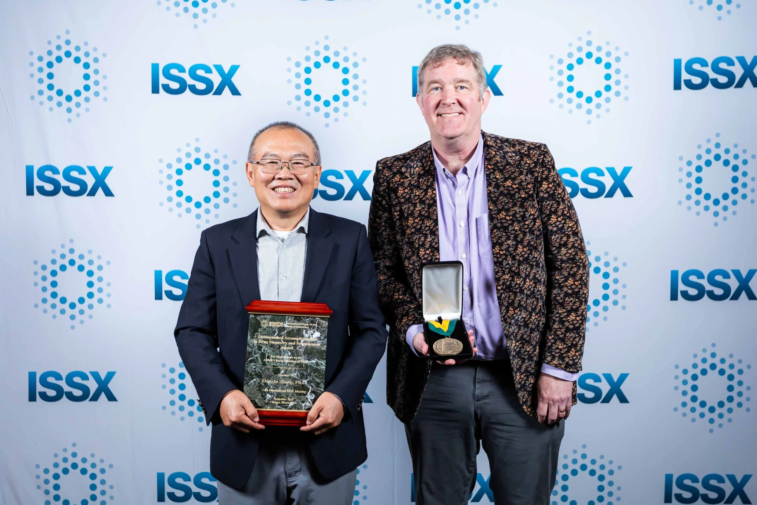 Two men standing in front of a white backdrop with ISSX logo, holding awards and smiling at the camera.
