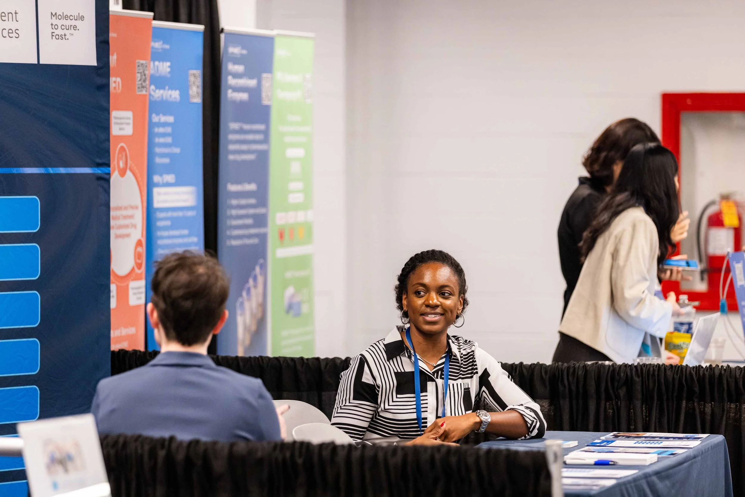 A woman with dark curly hair wearing a black and white striped shirt sitting at a booth talking to a man with brown hair wearing a gray suit. In the background, there are other people and informational posters, with a fire extinguisher on the wall.