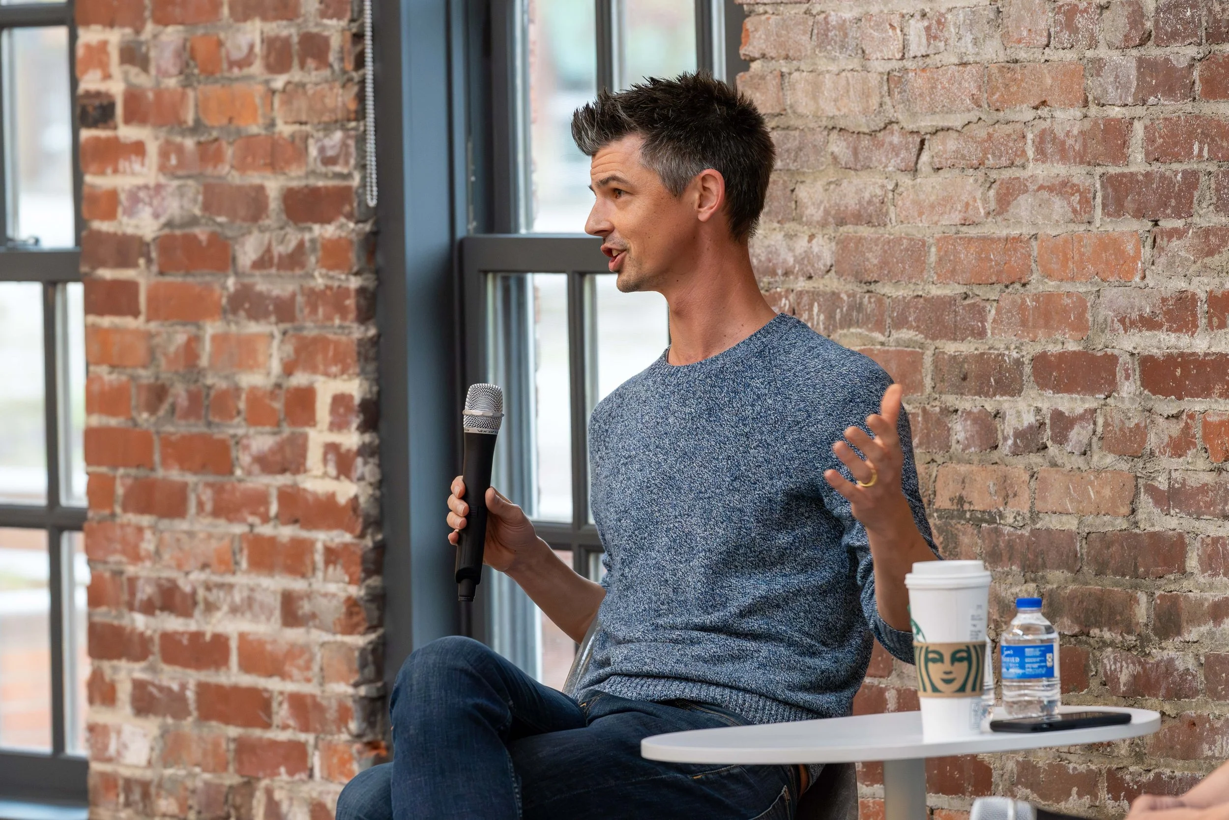 A man sitting in front of a brick wall, holding a microphone and speaking, with a coffee cup, bottle of water, and a phone on a small white table beside him.