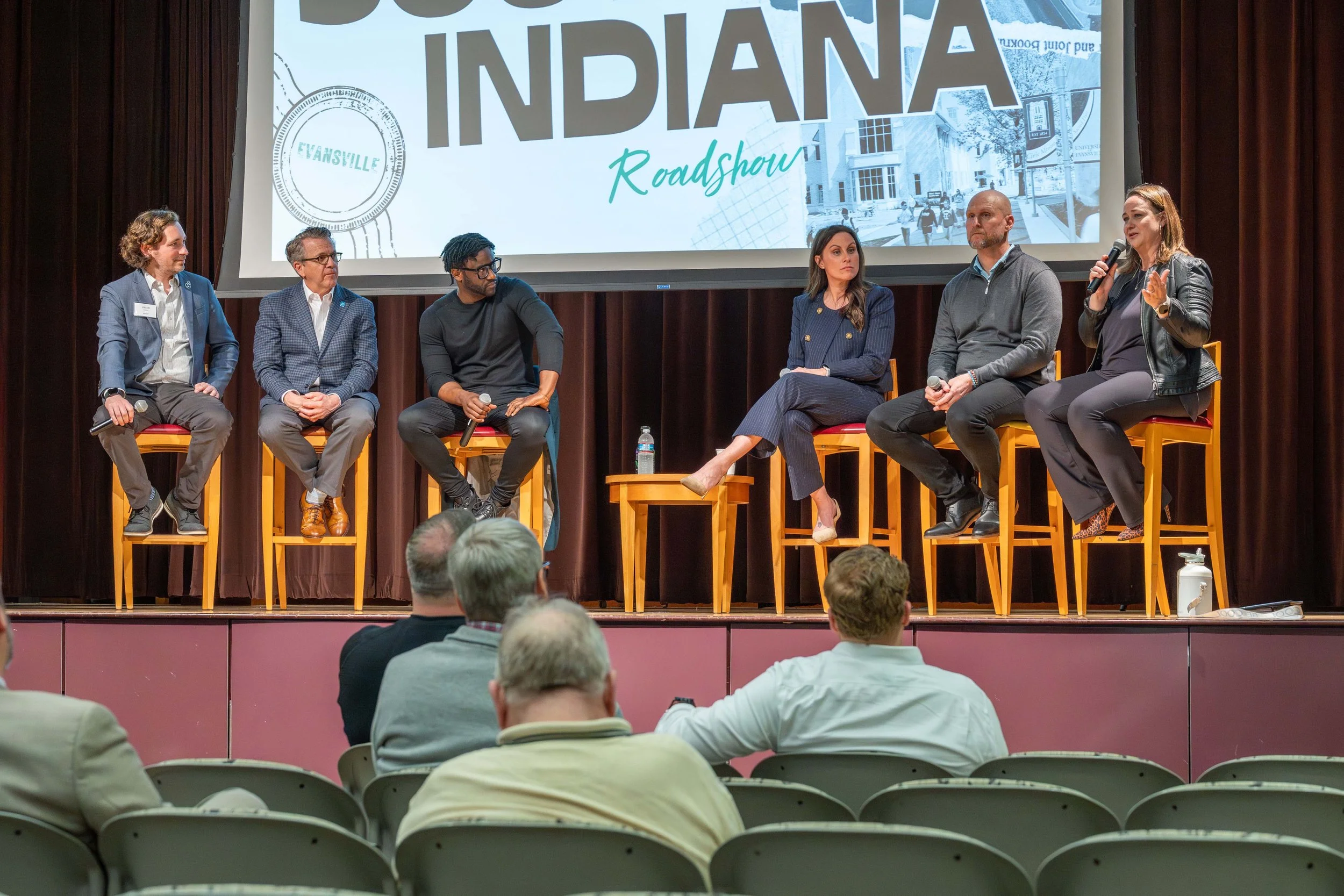 A panel discussion taking place on a stage with six people seated on chairs. A woman on the far right is speaking into a microphone. Behind them is a large screen displaying text that reads 'Indiana Roadshow' and a graphic of a cityscape. The audienc