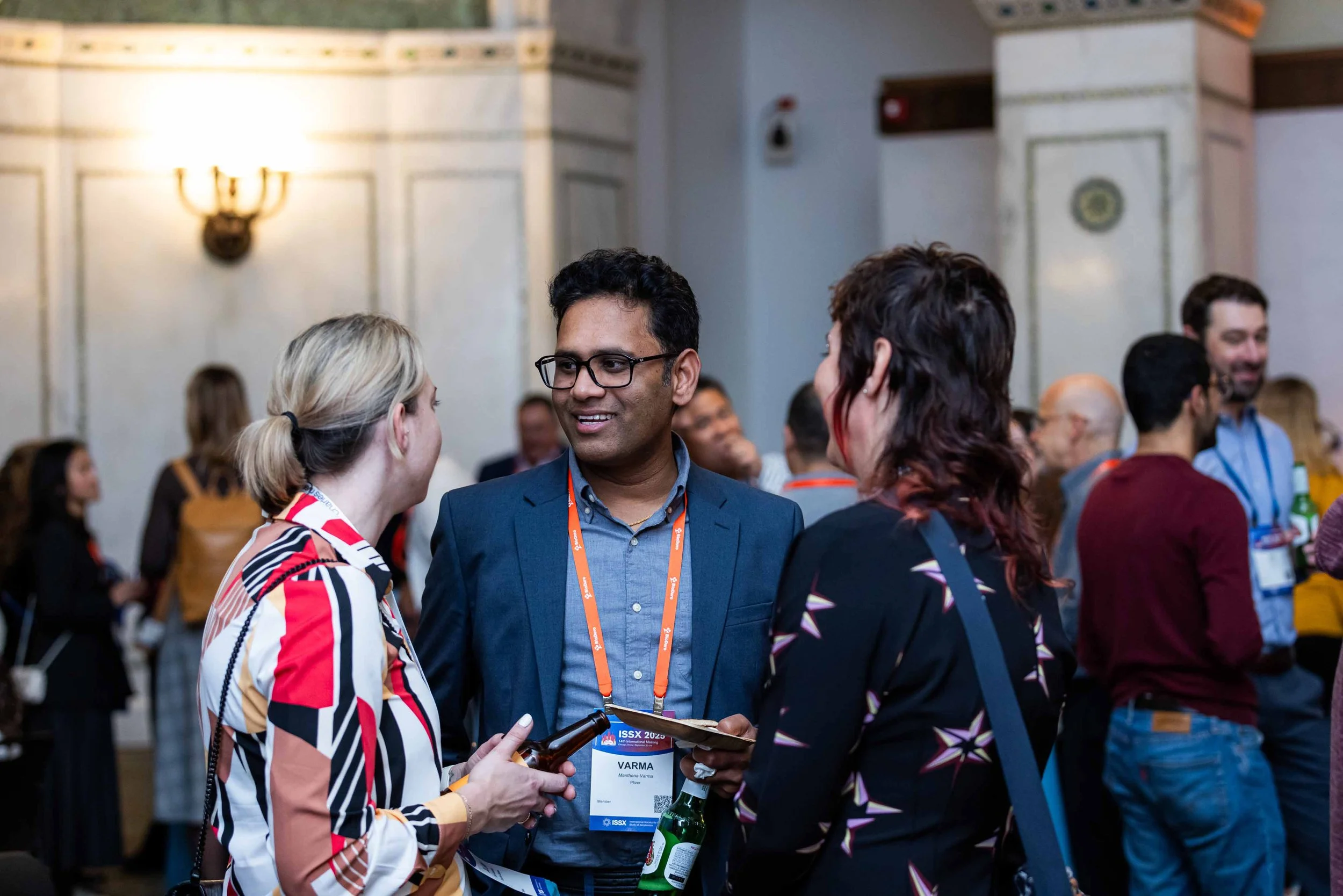 A group of people at a conference engaged in conversation, with a man in a blue suit and glasses smiling and talking to two women, all wearing conference badges, in a ballroom with ornate walls and other attendees in the background.