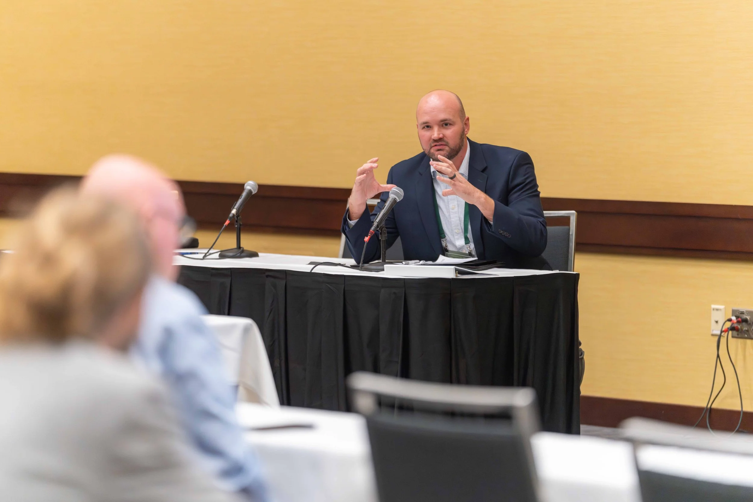 A man in a suit speaking at a conference, seated at a table with microphones, while an audience listens.