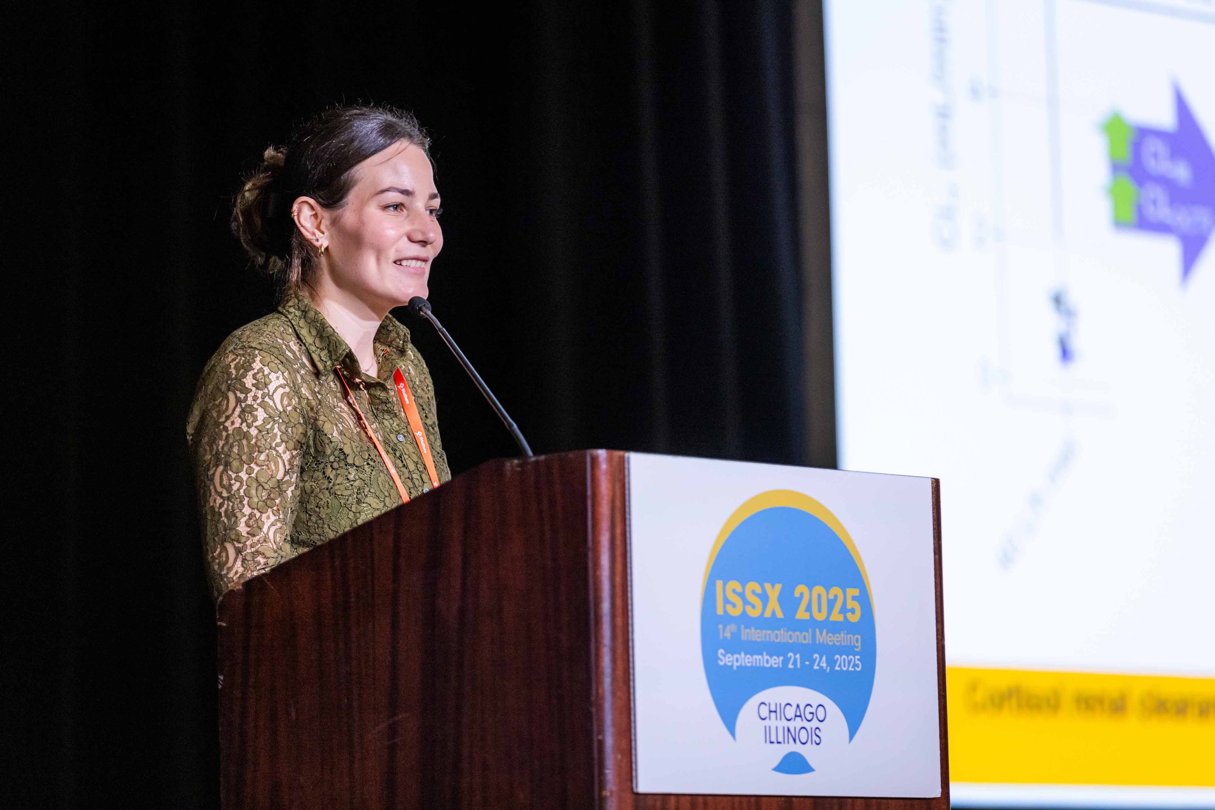 A woman is standing at a podium giving a presentation during the ISSX 2025 conference in Chicago, Illinois, from September 21 to 24, 2025. She is wearing a green lace top and smiling.