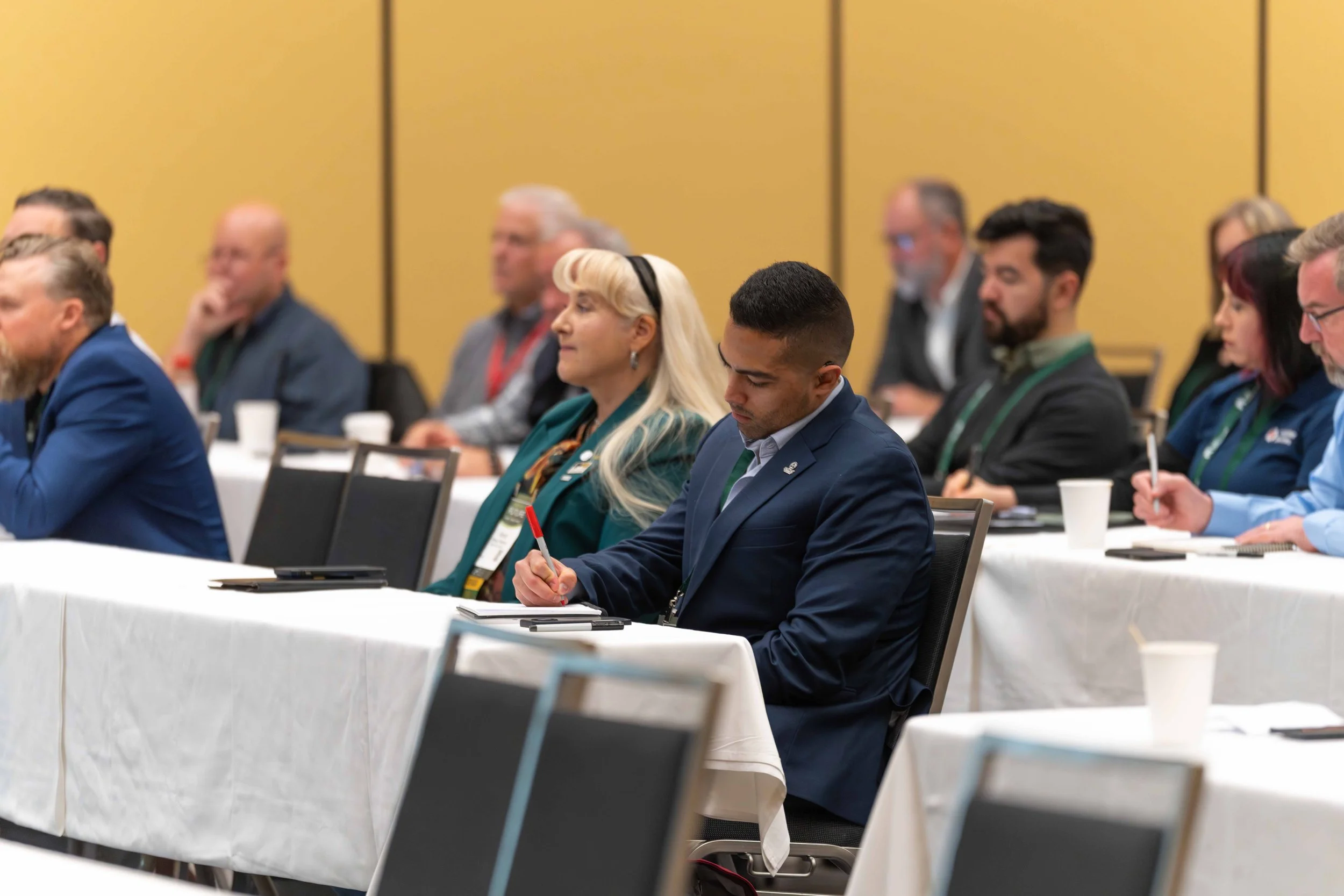People attending a conference or seminar, seated at tables with notepads and cups, listening attentively.