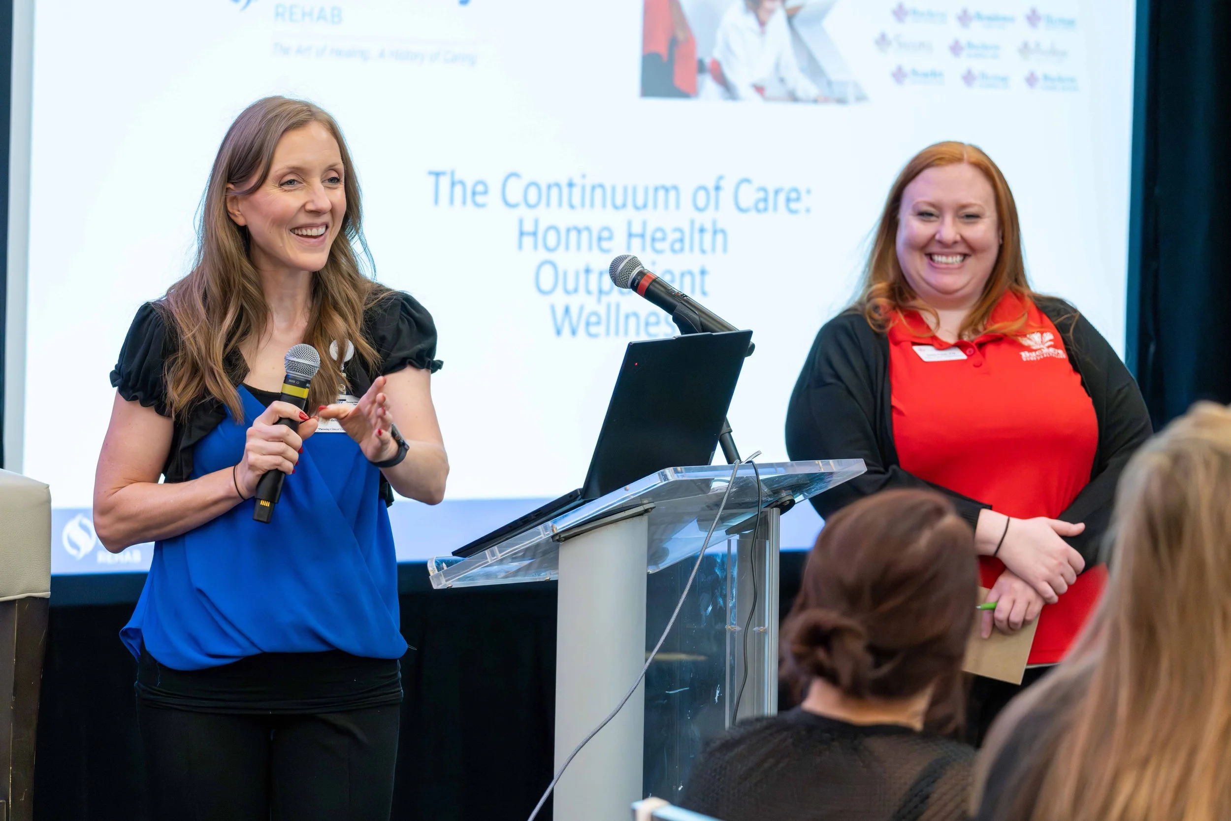 Two women speaking at a conference, one holding a microphone and smiling, the other standing beside her, with a presentation slide in the background titled 'The Continuum of Care: Home Health, Outpatient, Wellness'.