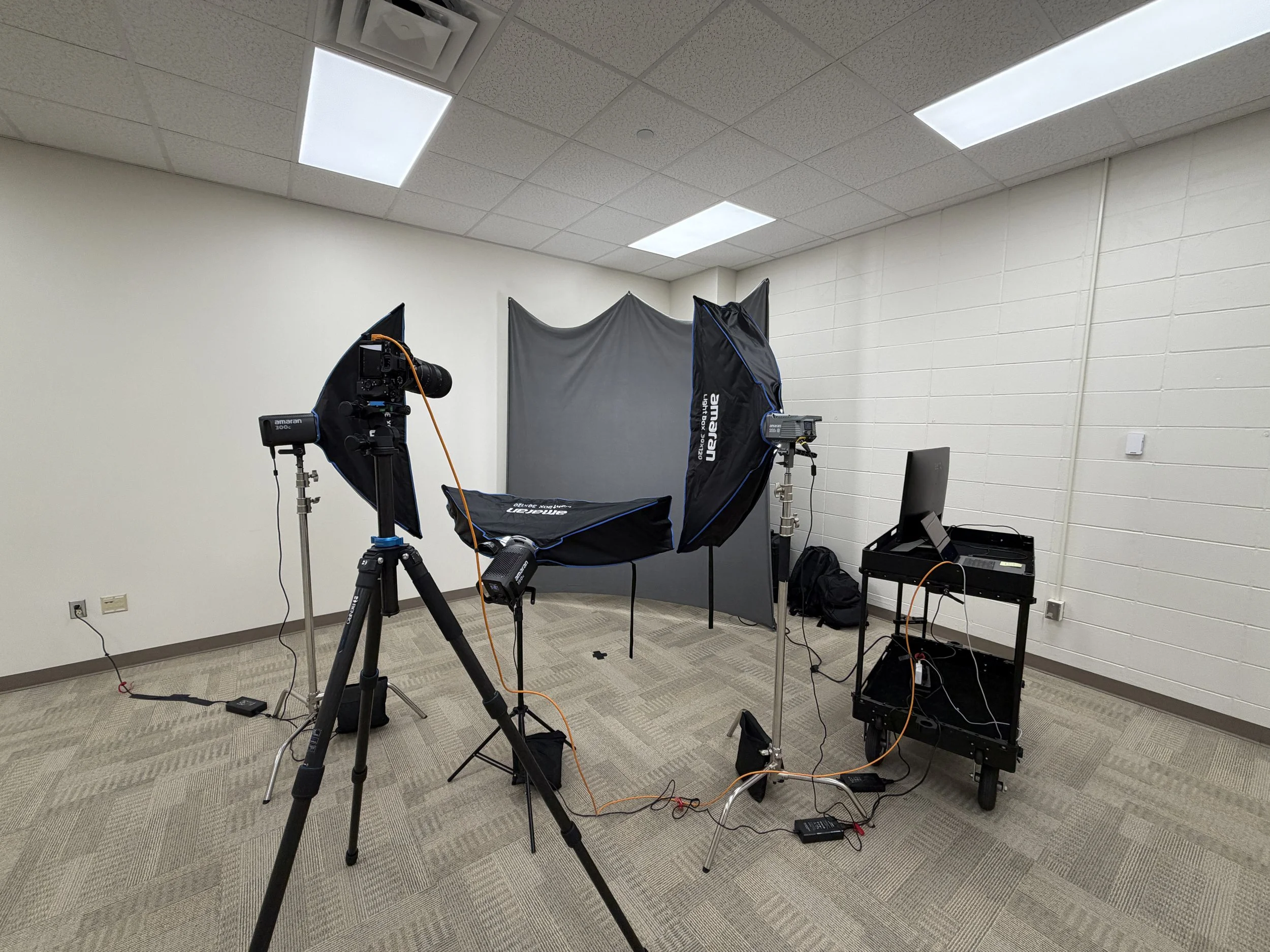 Photography studio setup with lighting equipment, a camera on a tripod, a backdrop, a computer, and related accessories in an empty room.