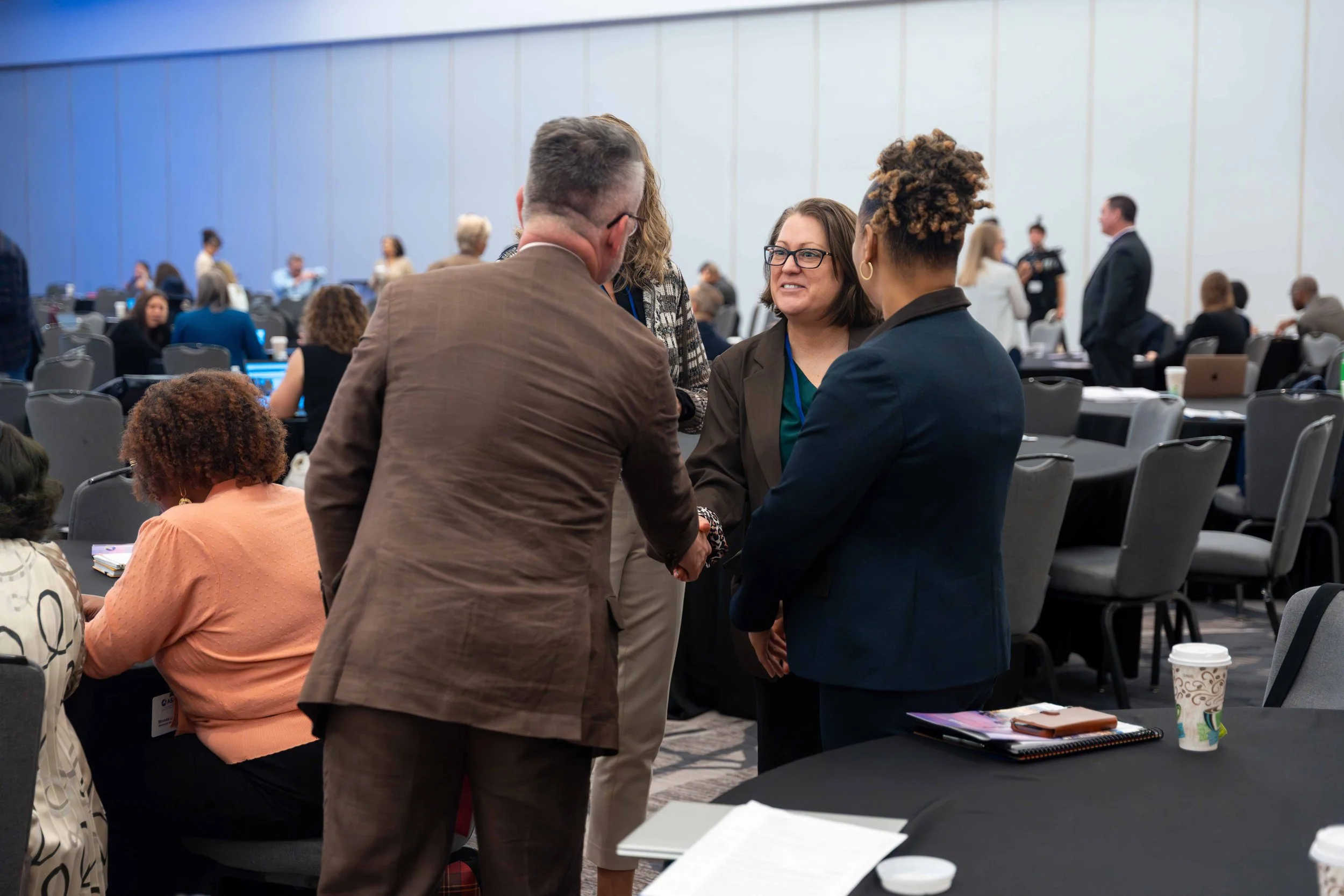 People in a conference room engaging in conversation, with chairs and tables filled with papers and cups, some working on laptops.