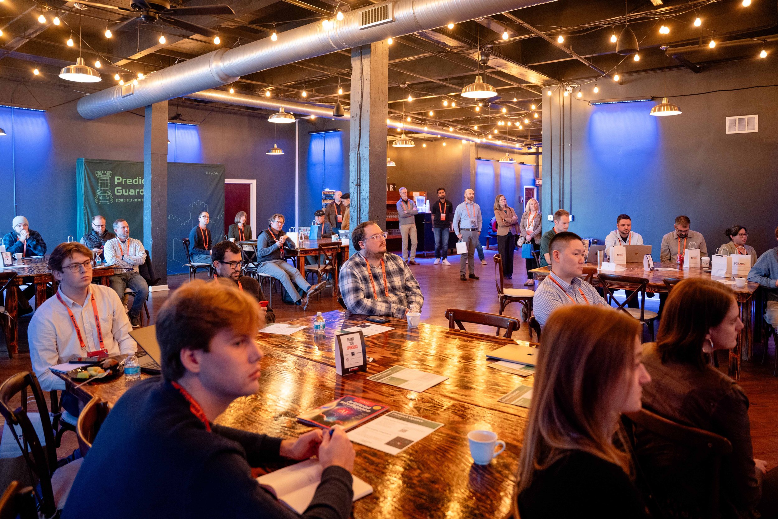 People attending a conference in a dimly lit room with string lights and blue curtains, seated at tables and listening to a speaker.