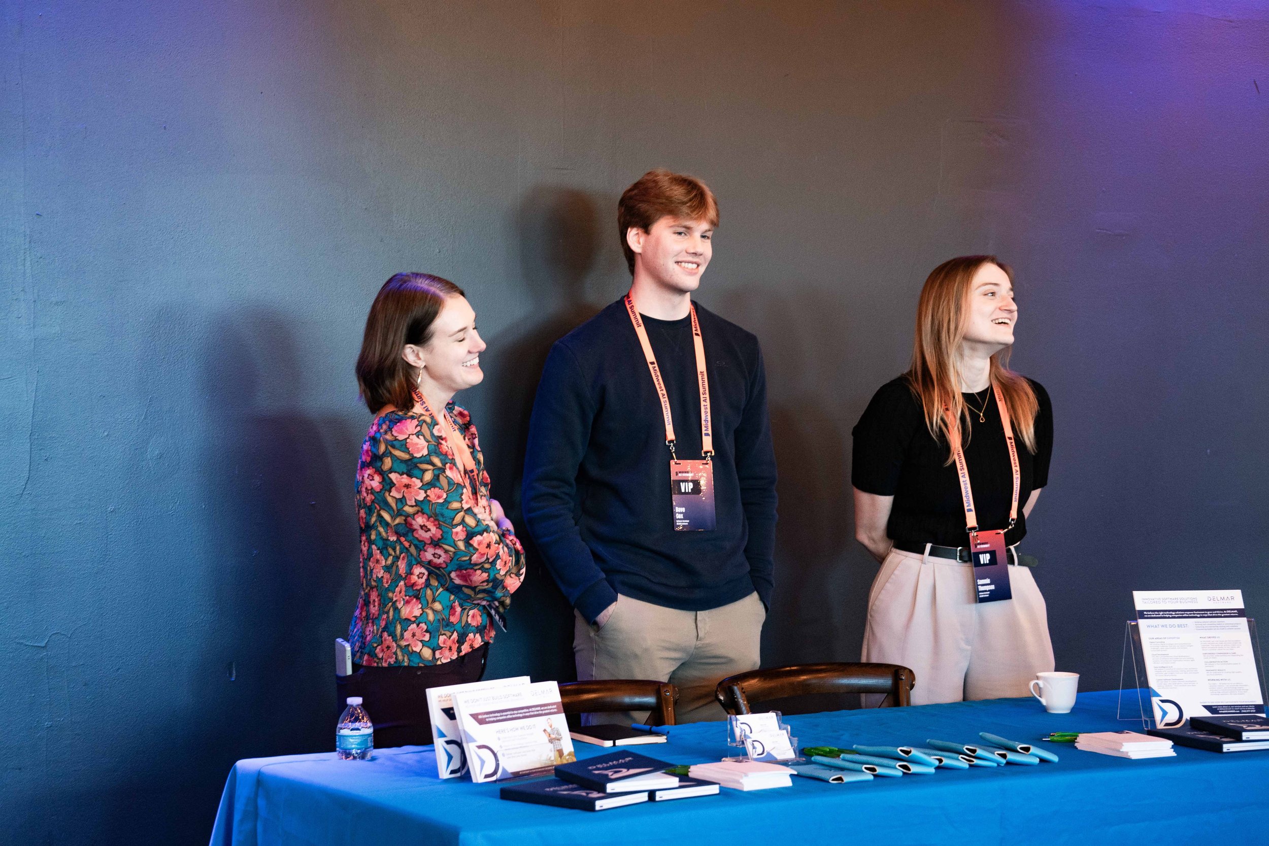 Three people standing behind a conference registration table, smiling and looking to the side, with conference badges hanging from their necks.