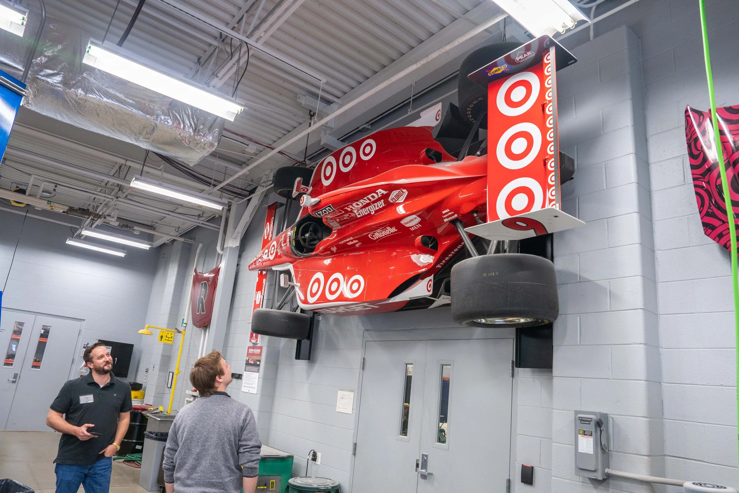 A red race car with sponsor decals mounted on a wall inside a garage. Two men are standing in front, looking at the car.
