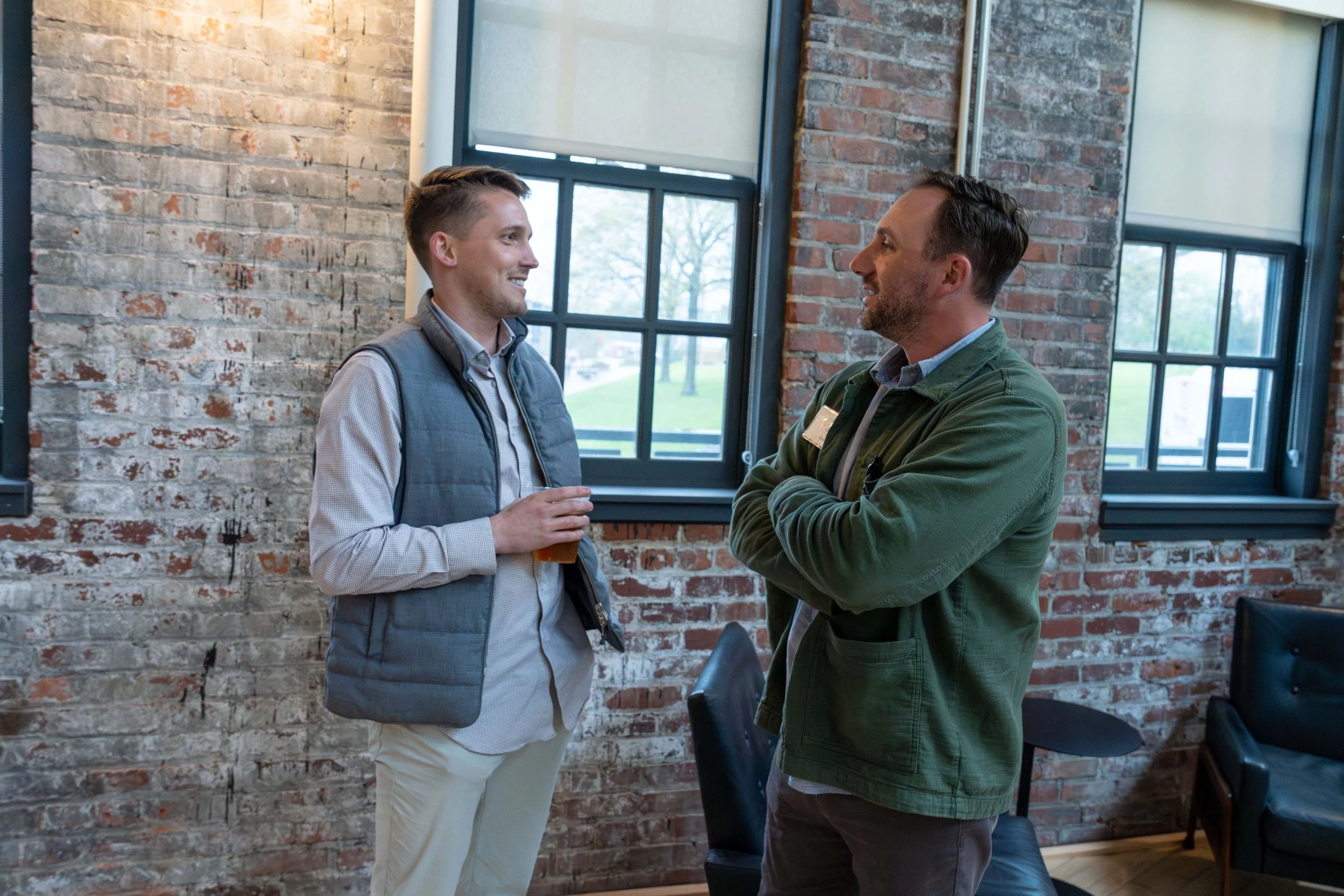 Two men having a conversation in a room with exposed brick walls and large windows, one holding a drink.