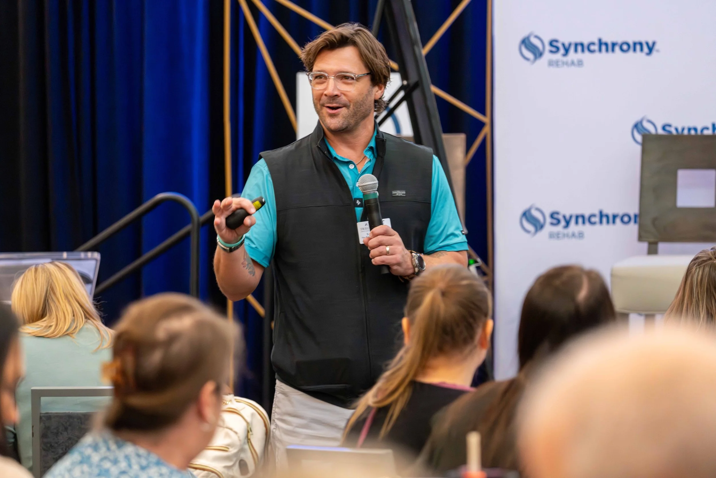 A man giving a presentation at a conference, holding a microphone and a remote, with an audience watching. There are banners with the 'Synchrony' logo in the background.