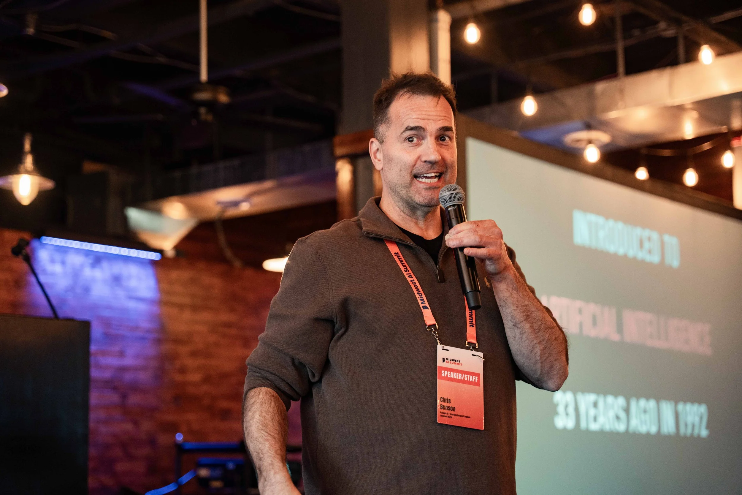 A man speaking into a microphone at an indoor event, wearing a brown sweater and a red conference badge with his name, Chris Benson, around his neck. In the background, there is a large screen displaying text and a brick wall.