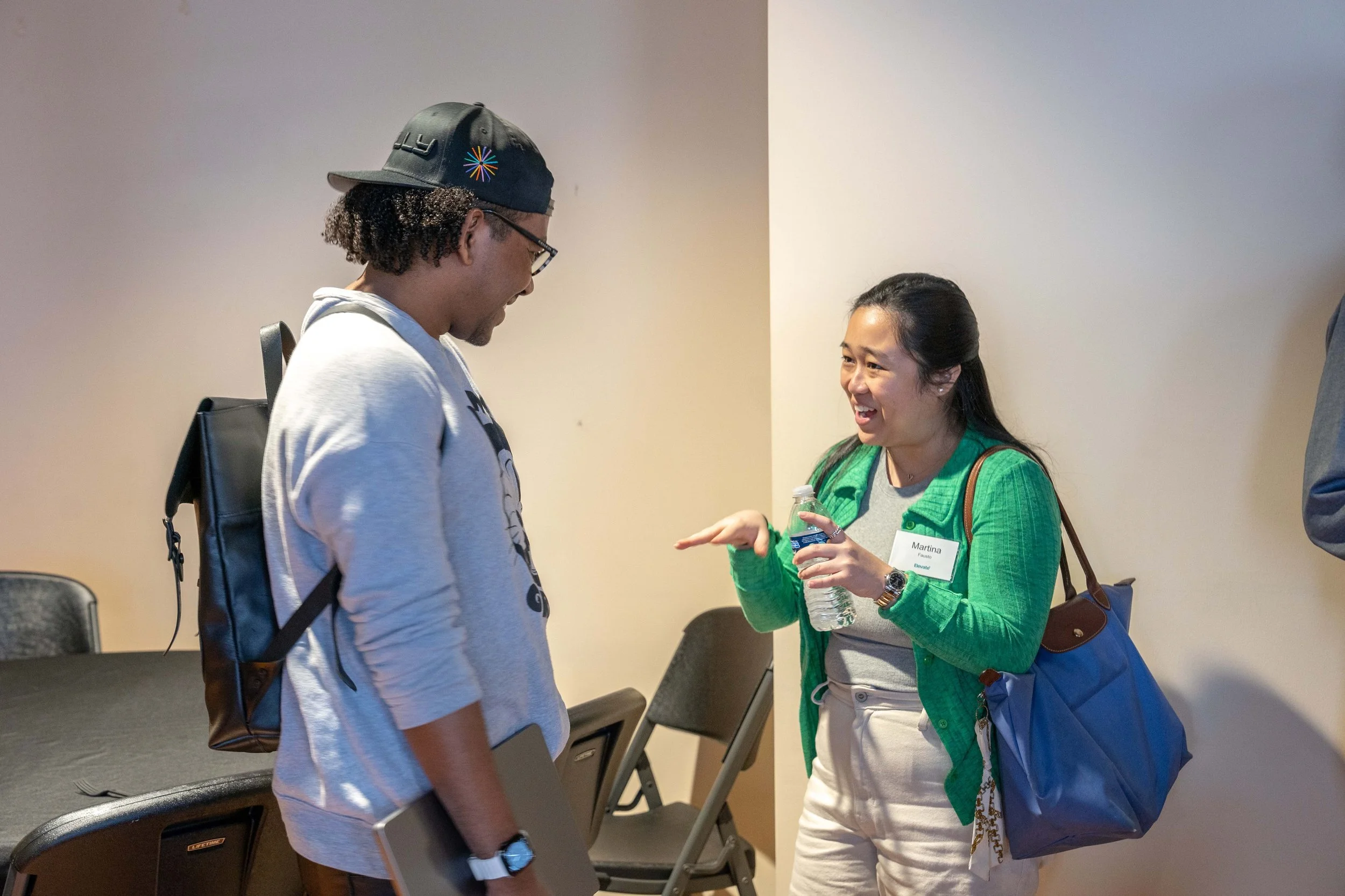 A woman in a green jacket and gray shirt smiling and talking to a man wearing a gray hoodie, glasses, and a baseball cap indoors.