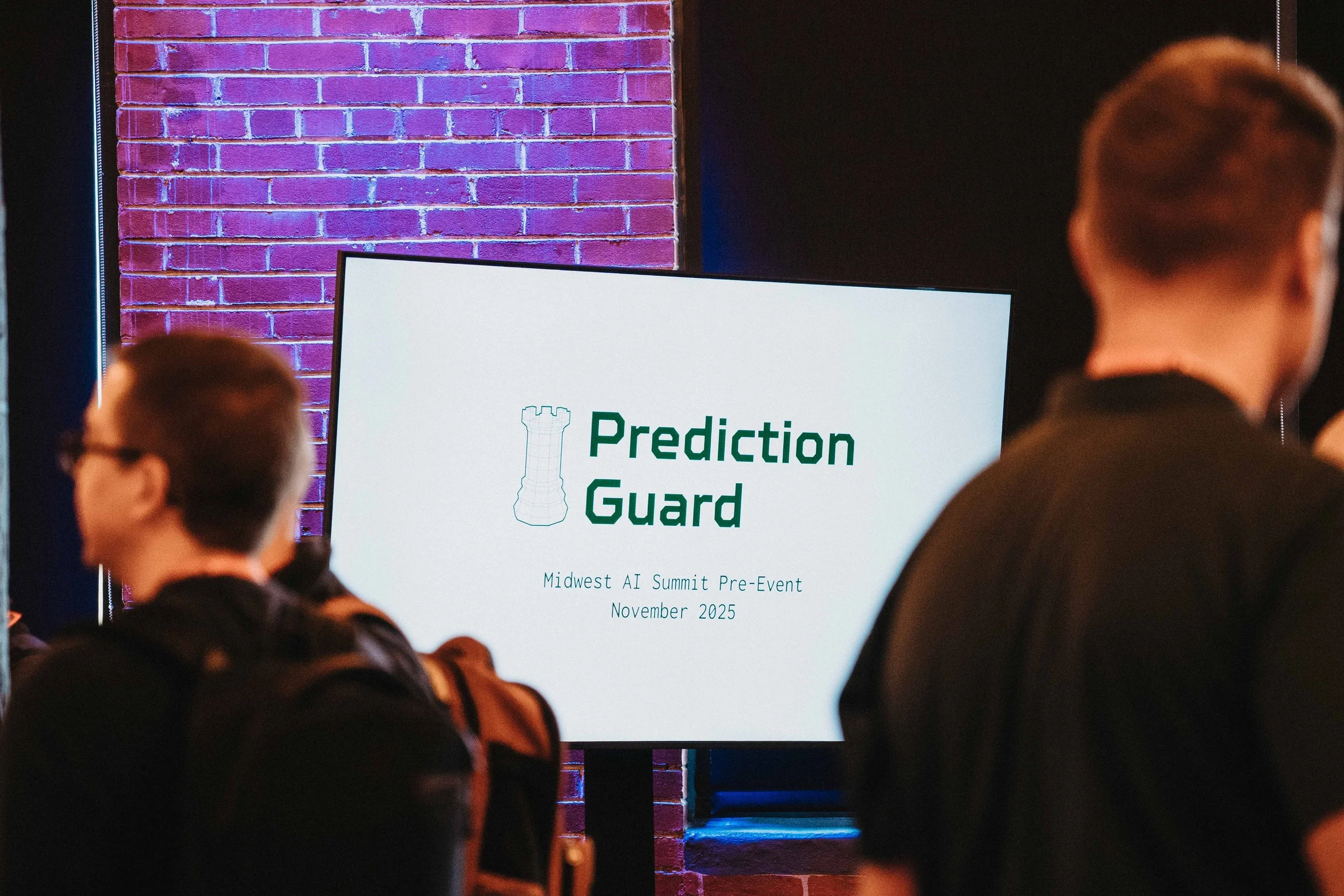 A digital screen displays the  logo and event information for 'Prediction Guard' at the Midwest AI Summit Pre-Event in November 2025, with people in the foreground attending the event.