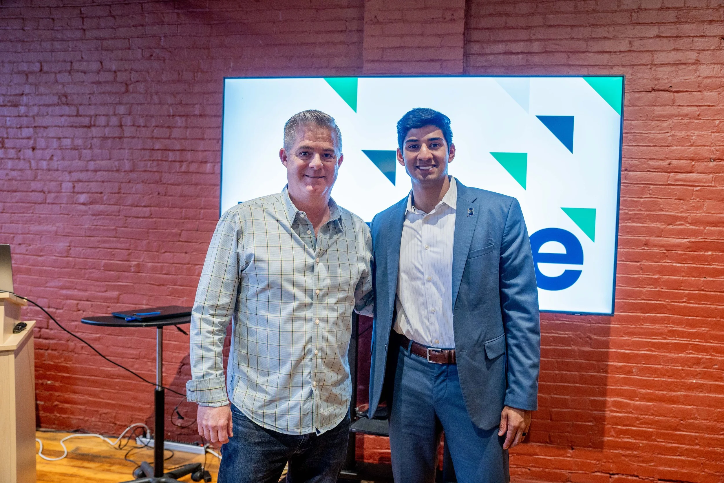 Two men standing side by side in front of a digital screen with a geometric design and the text 'e'. The man on the left is wearing a plaid button-up shirt and jeans, with short gray hair. The man on the right is wearing a suit with a pin on his lape