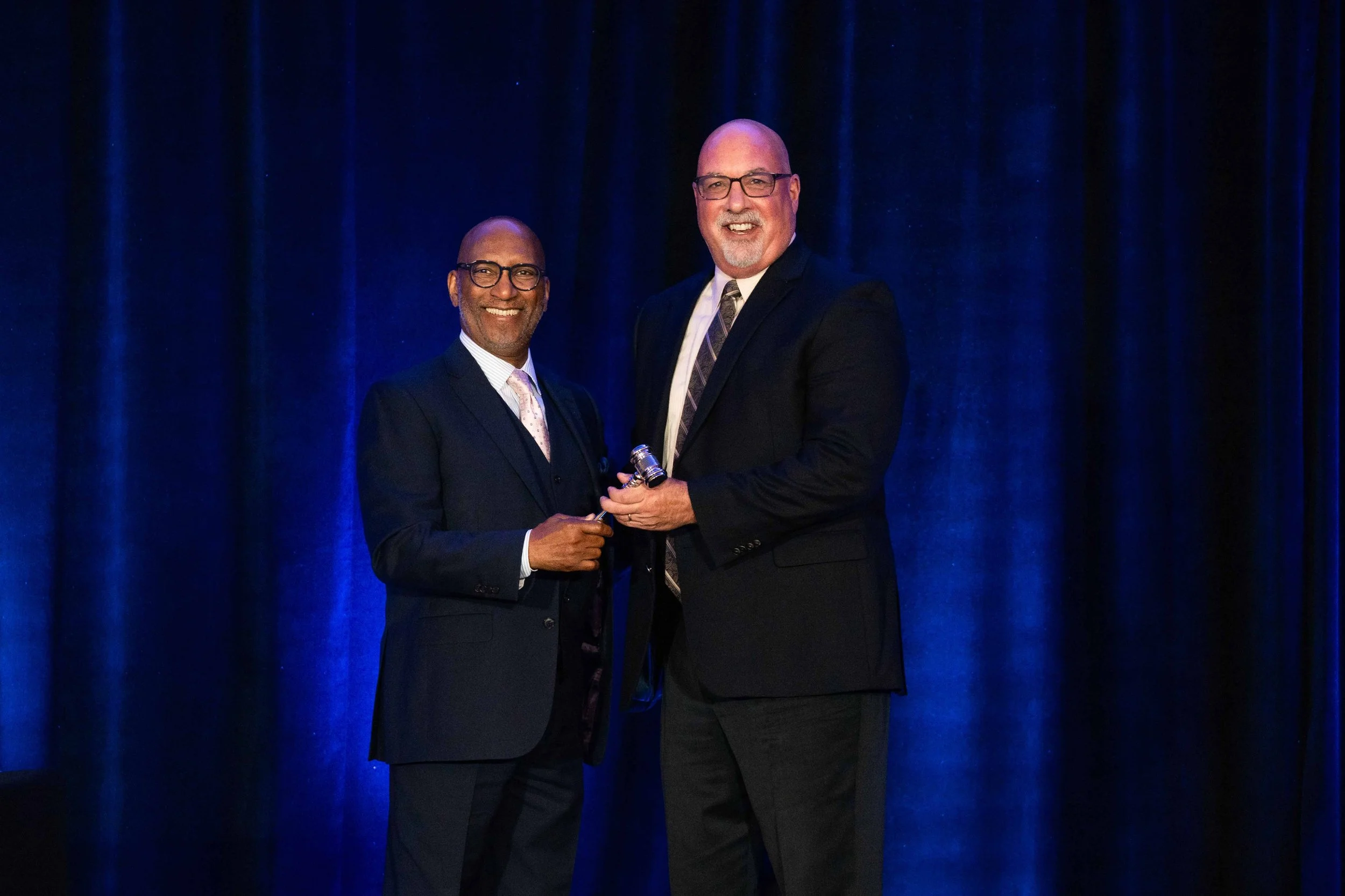 Two men in suits on a stage, one handing an award to the other, with dark blue curtains in the background.