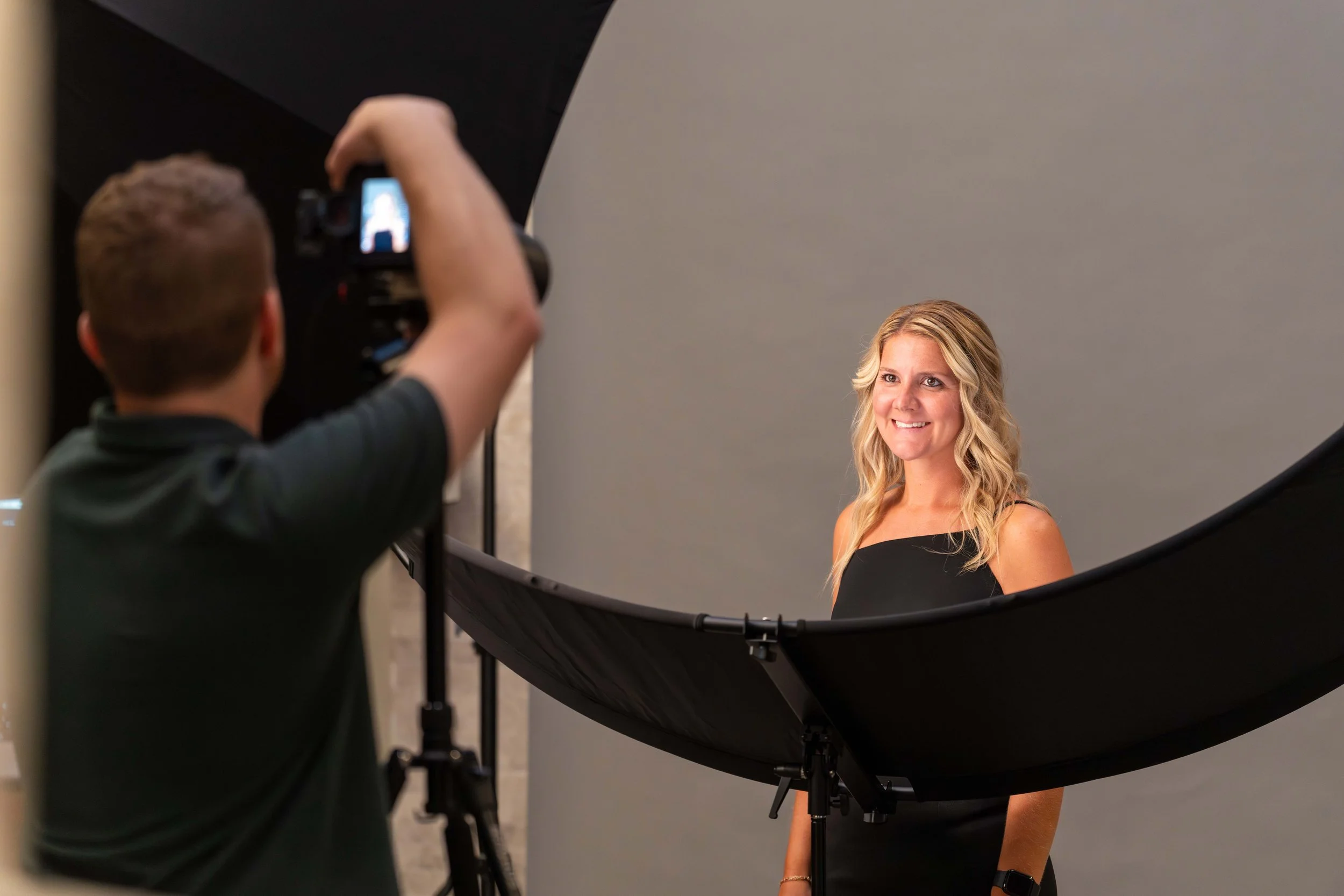 Photographer taking a portrait photo of a smiling woman in front of a gray backdrop.