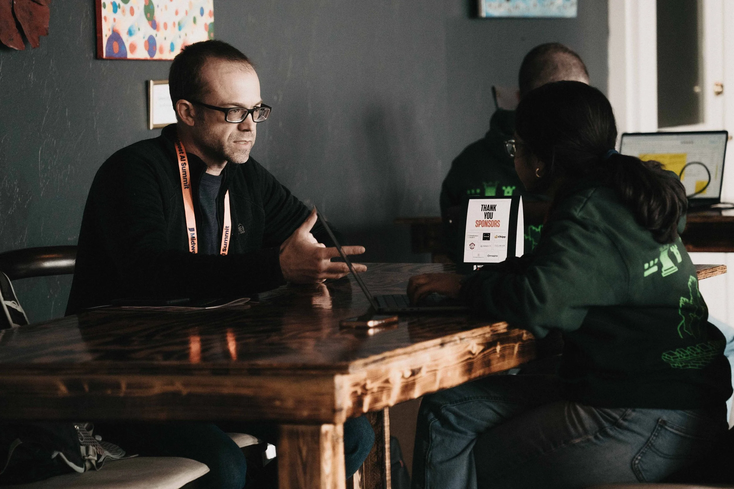 Three people sitting at a wooden table, engaged in a conversation with one using a laptop. The setting appears to be indoors with abstract artwork on the wall.
