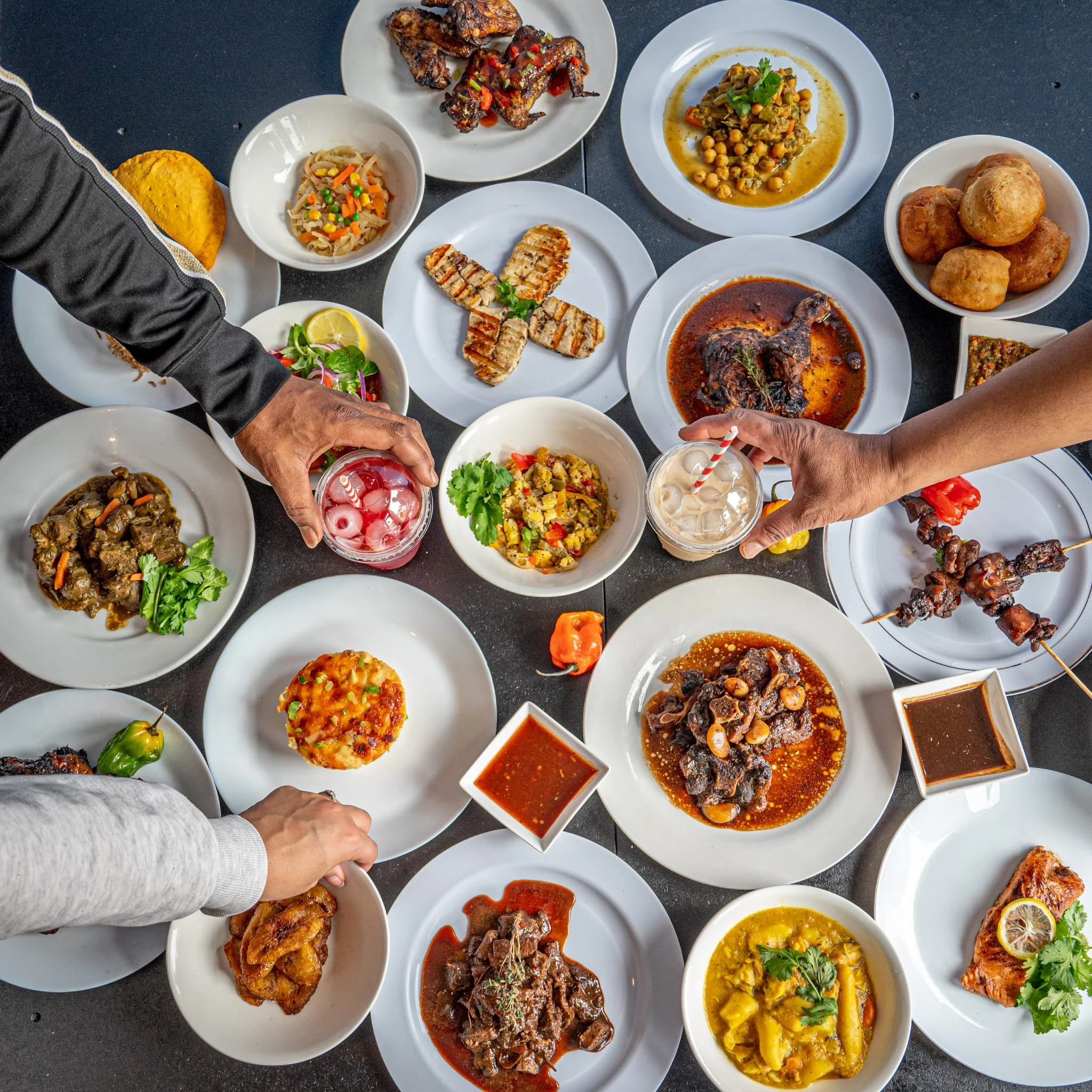 Assorted Caribbean dishes on a table, including grilled meats, stews, plantains, and beverages, with people reaching for the food.
