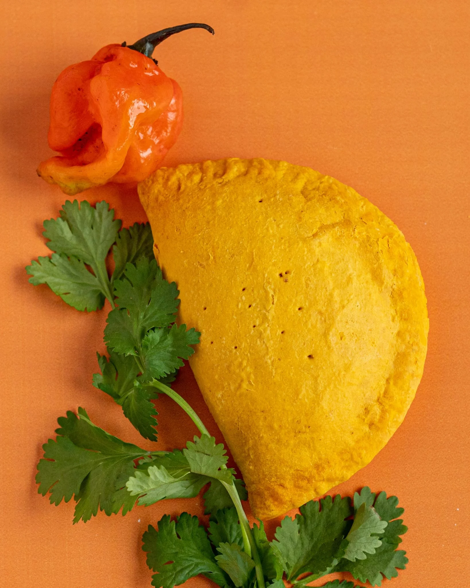 A Jamaican patty with a Scotch bonnet pepper and cilantro on an orange background.