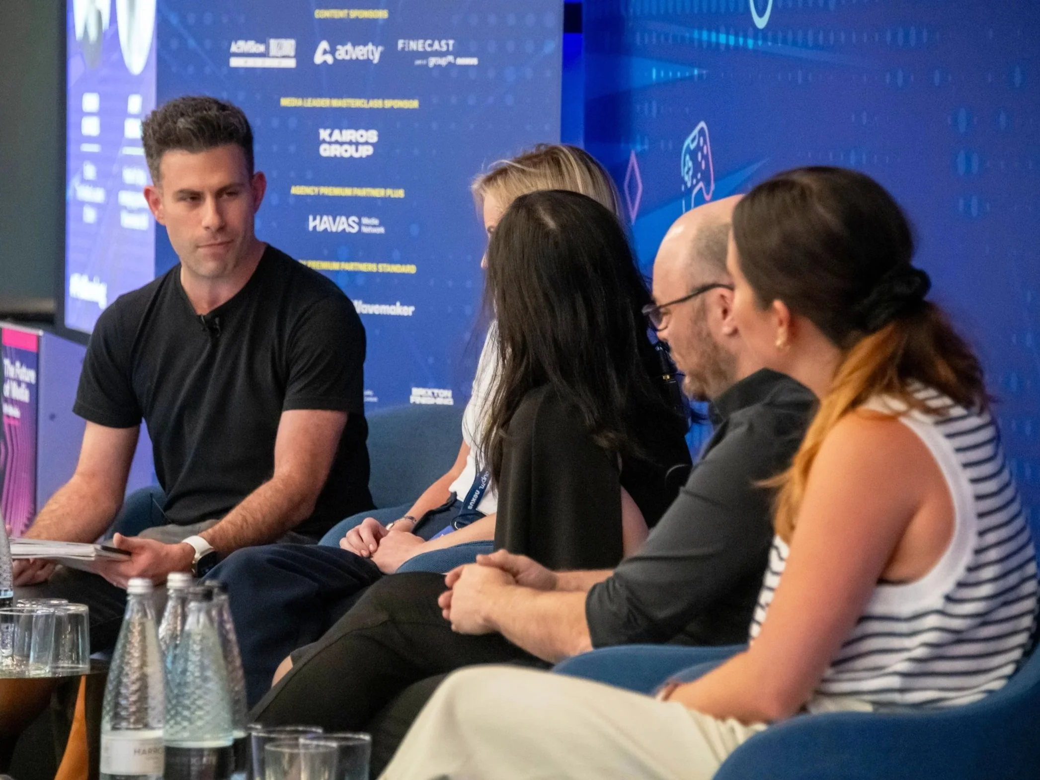 People sitting at a panel discussion or conference, with a blue backdrop showing various logos and text, and water bottles on the table in front of them.