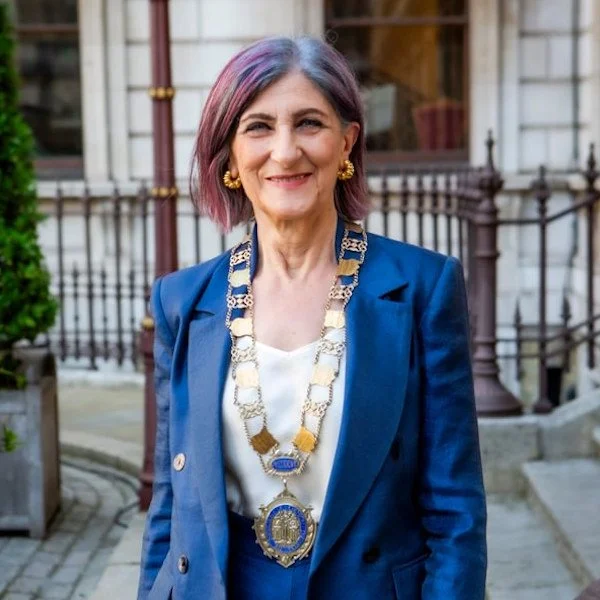 A smiling woman with shoulder-length, multi-colored hair, wearing a blue blazer, a white blouse, and a large statement necklace with gold and blue details, standing outdoors in front of a building with a wrought iron fence.