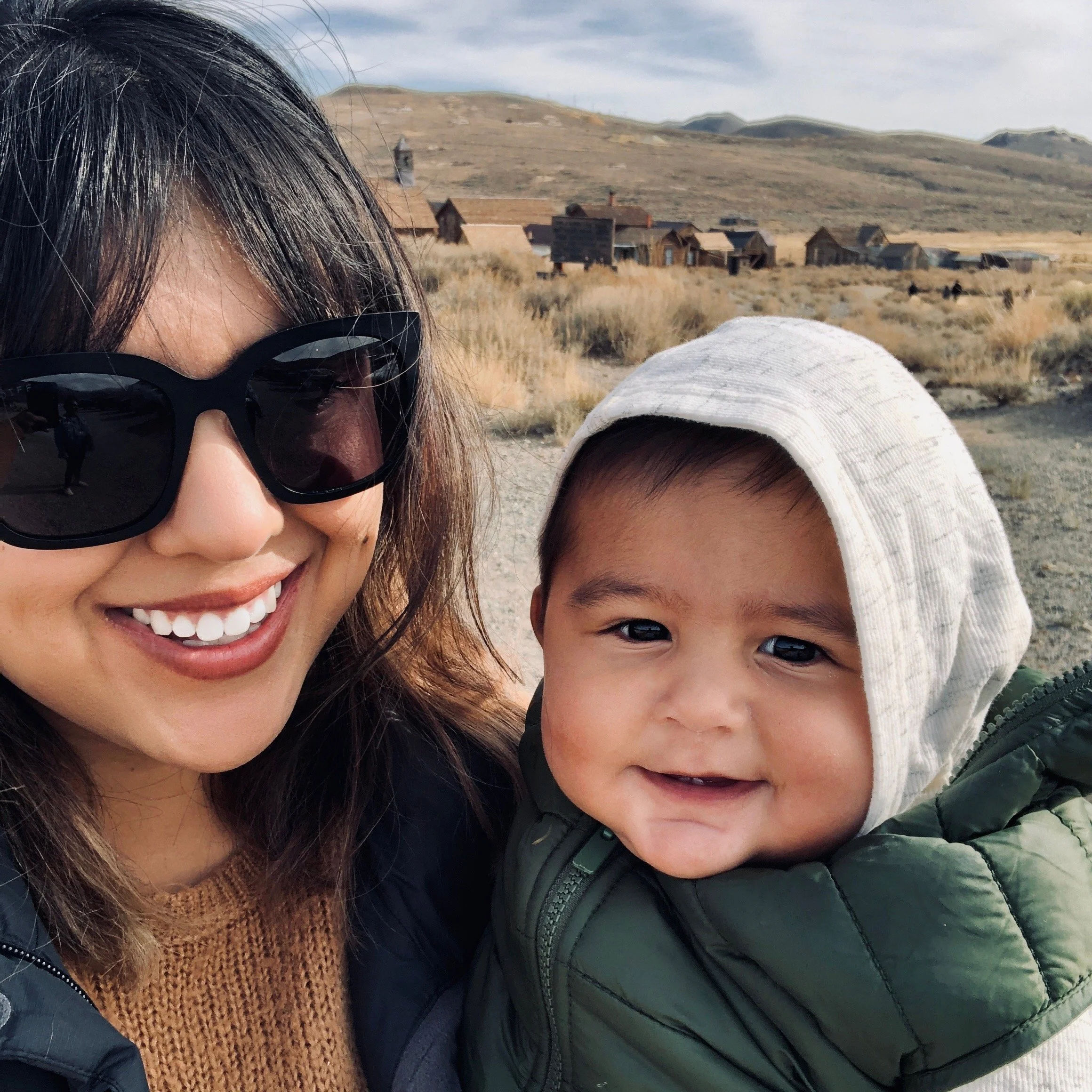 A woman with long brown hair wearing large black sunglasses smiling next to a young child with short dark hair, a light-colored hoodie, and a green jacket outdoors in a rural area with dry grass, hills, and old wooden buildings in the background.