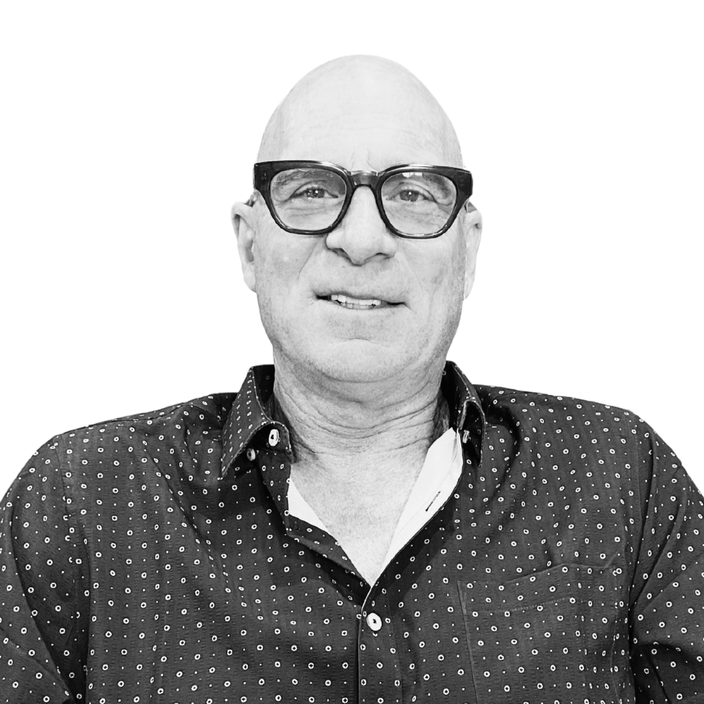 A black and white photo of a bald man wearing large black glasses and a dark button-up shirt with small white patterns, smiling slightly.