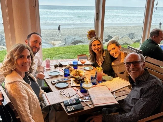 Group of five people sitting at a table in a restaurant with large windows overlooking a beach and ocean, with drinks and menus on the table.