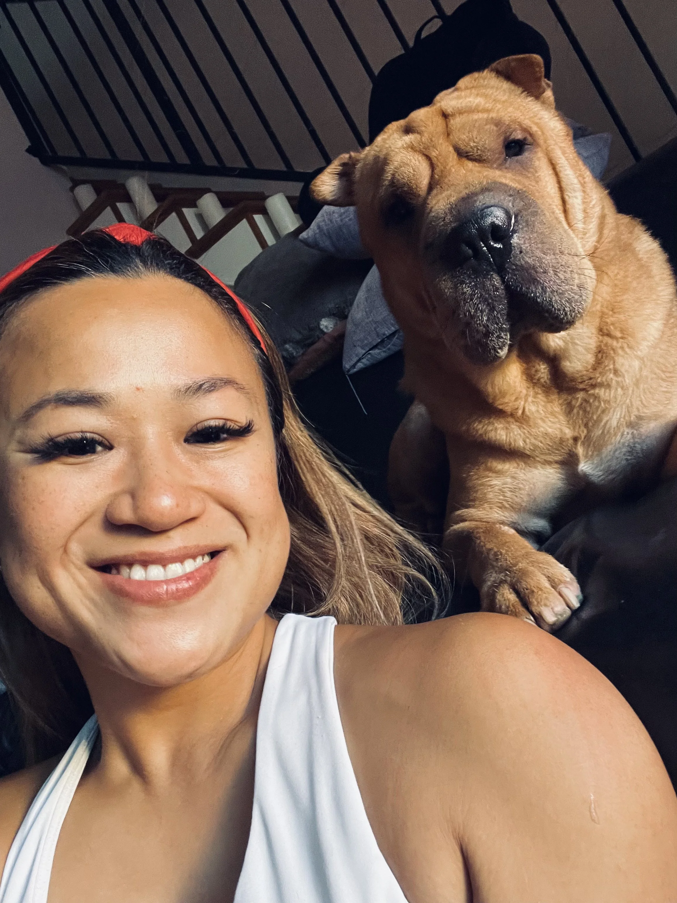 A woman smiling with a large brown dog with a wrinkled face sitting next to her indoors.