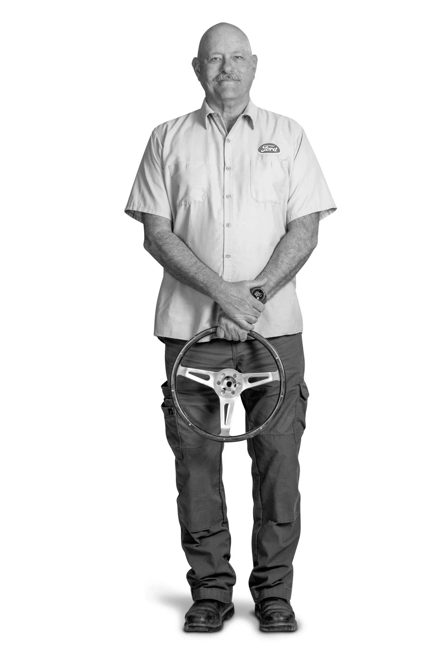 A man dressed in a short-sleeved shirt with a Ford logo, dark pants, and boots, holding a steering wheel, standing against a plain background.