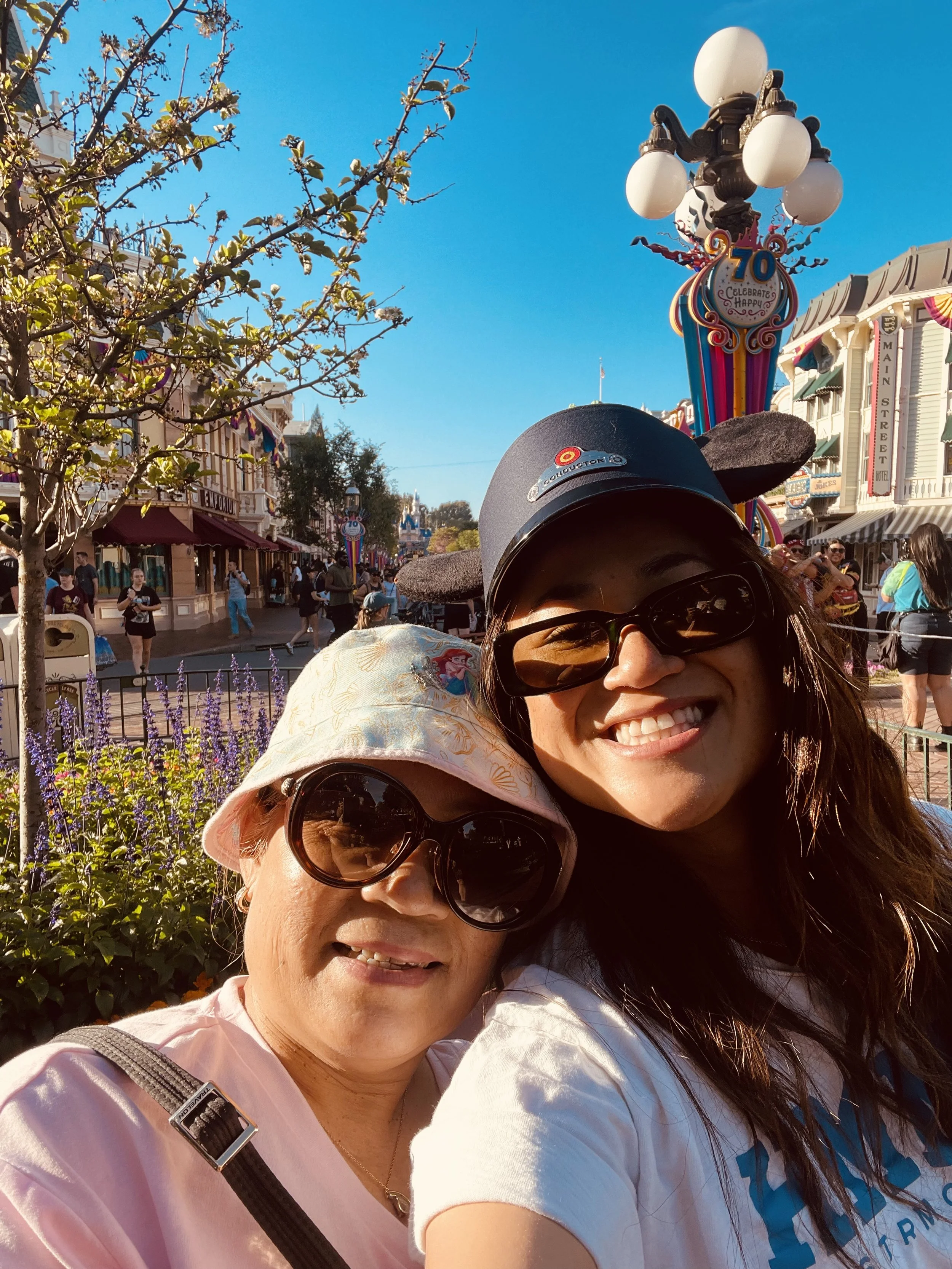Two women smiling and taking a selfie at Disneyland, with a colorful lamp post celebrating the park's 70th anniversary in the background.
