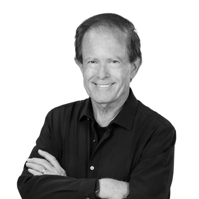 Black and white portrait of a smiling middle-aged man with short, slightly wavy hair, wearing a dark, long-sleeved, collared shirt with arms crossed against a plain white background.