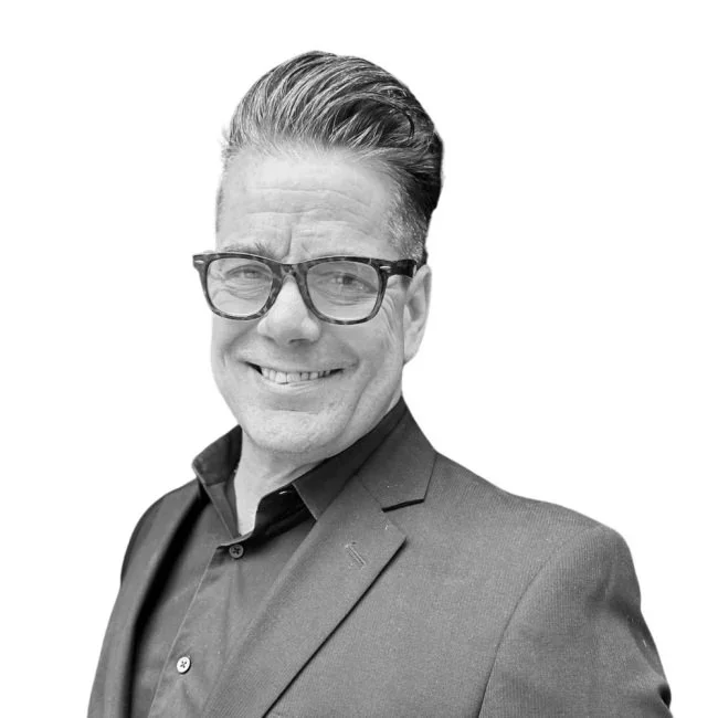 A man with glasses and neatly combed hair, wearing a dark suit jacket and a collared shirt, smiling at the camera against a plain background.