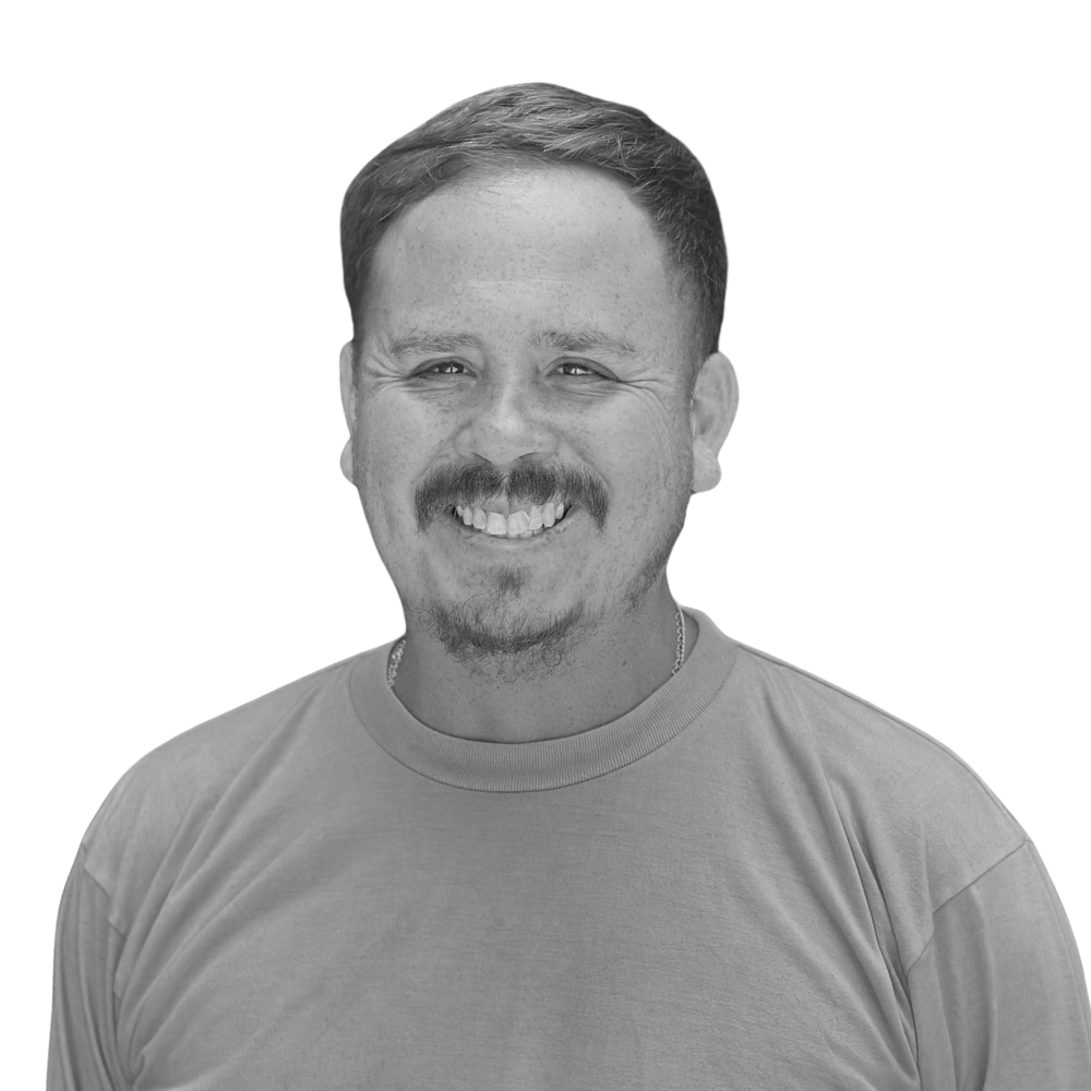 A man with a mustache and short hair smiling, wearing a t-shirt, in a black and white photo.