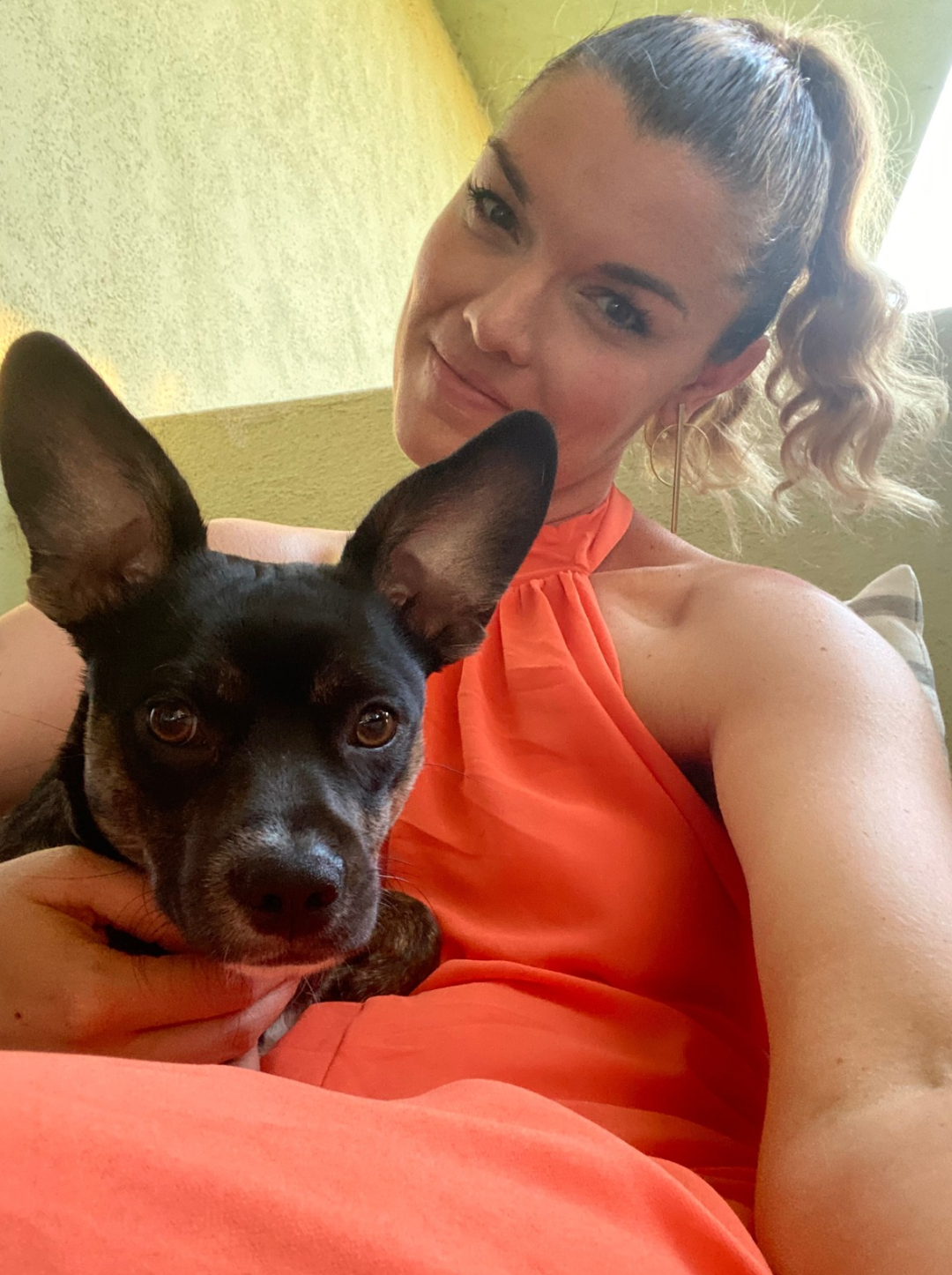 A woman with blonde, curly hair tied in a high ponytail, wearing an orange sleeveless top, taking a selfie with a small black and brown dog with large ears, sitting on her lap.