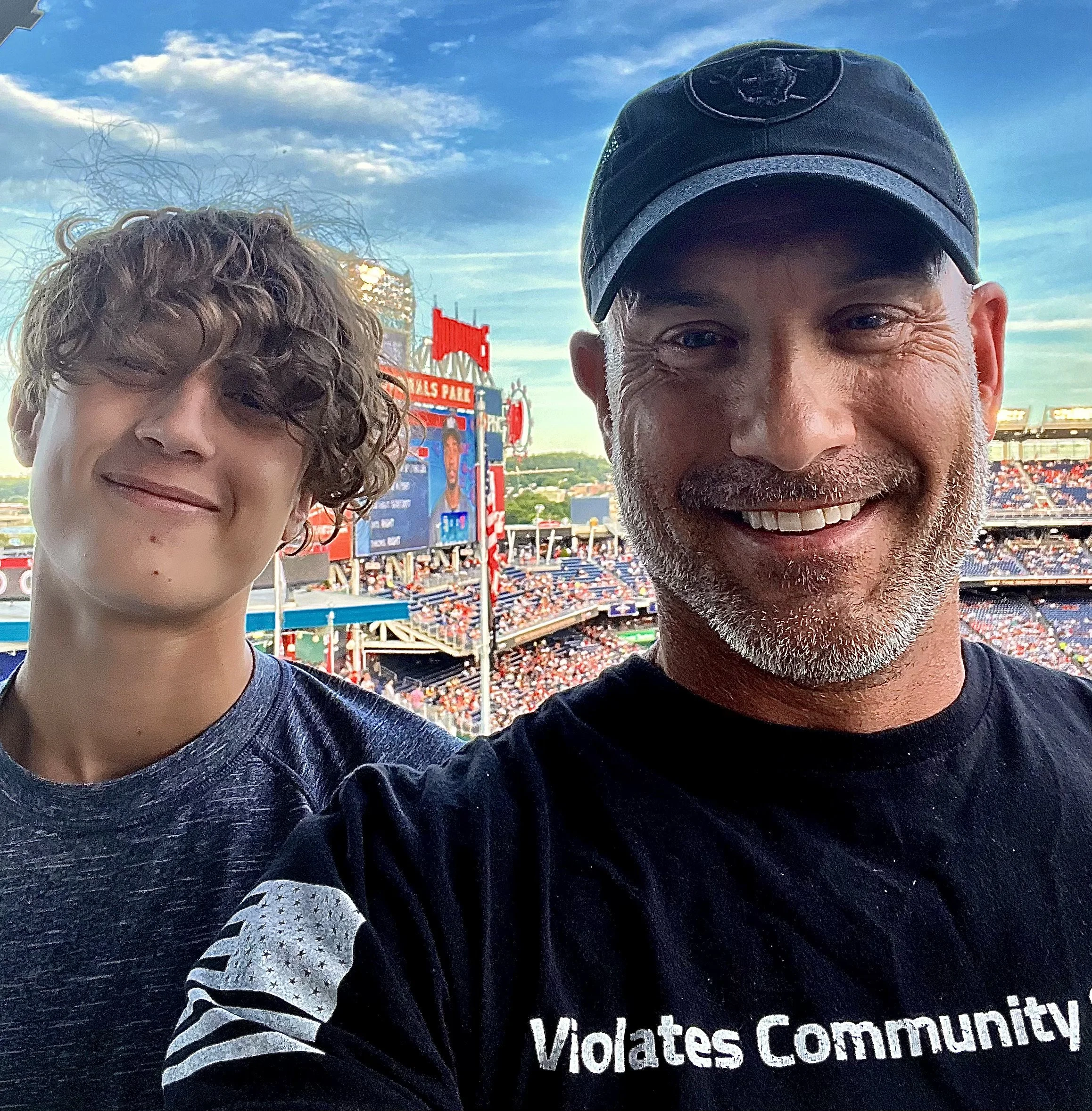 Two men taking a selfie at a baseball stadium during sunset, with stadium seating and a scoreboard in the background.