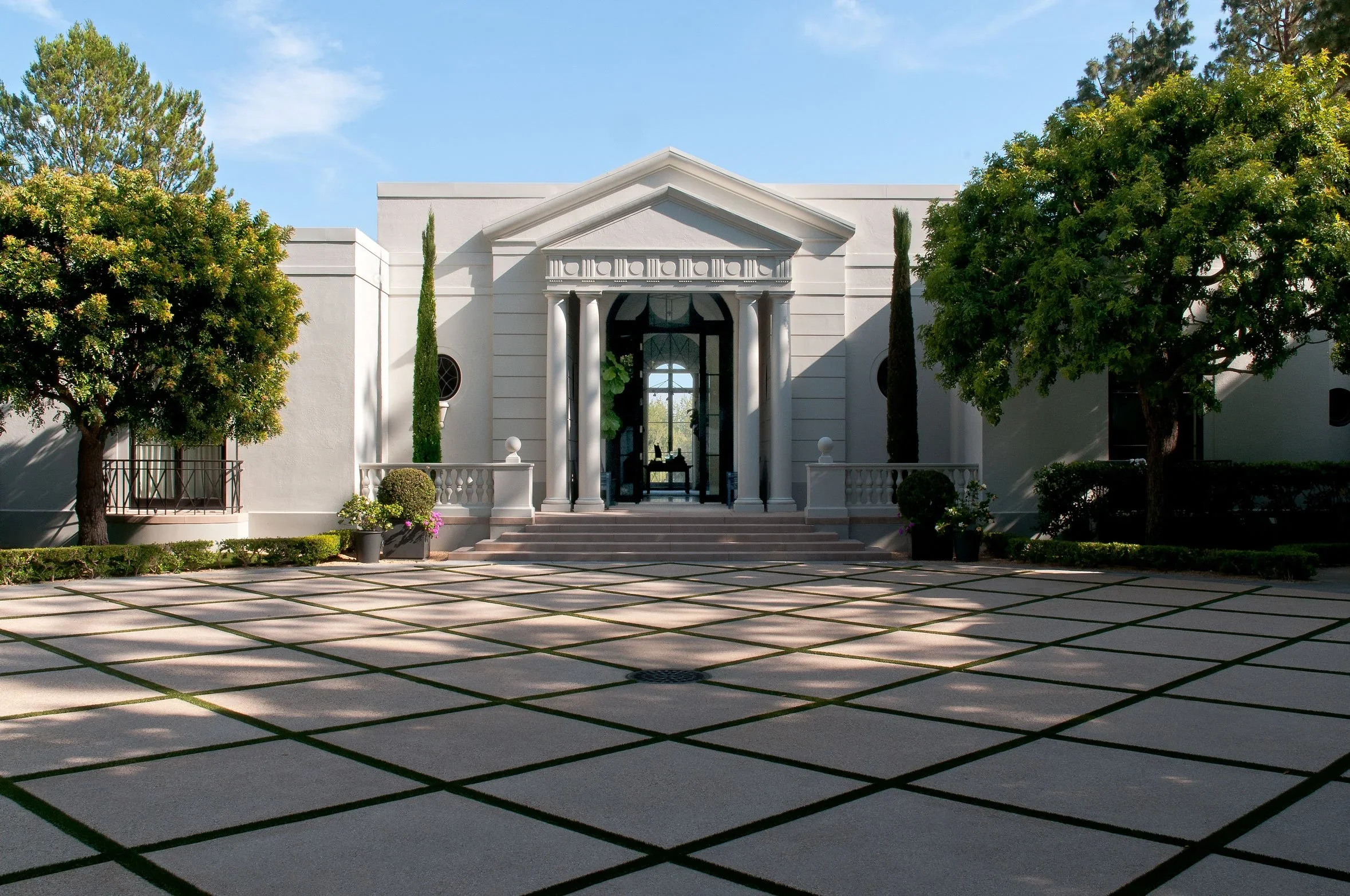 Exterior view of a white mansion with classical columns, stairs, and a symmetrical design, framed by trees on each side, with a tiled courtyard in the foreground under a blue sky.