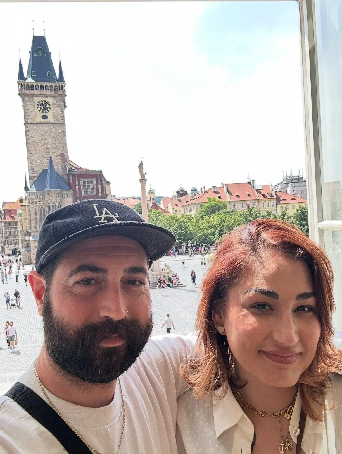 A man with a beard wearing a dark baseball cap and a woman with reddish-brown hair smiling in front of a window with a city square and a clock tower in the background.