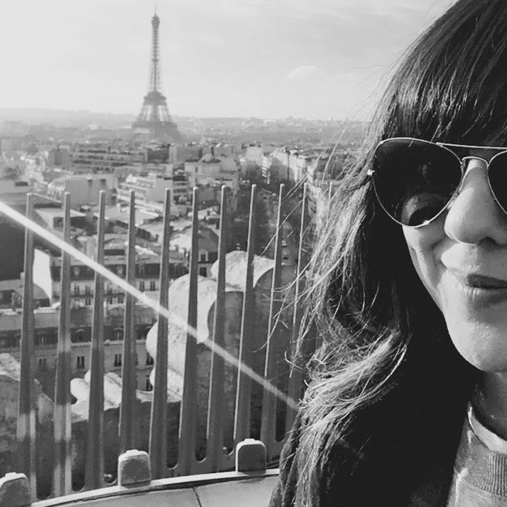 A woman with dark wavy hair wearing sunglasses, taking a selfie with the Eiffel Tower in the background.