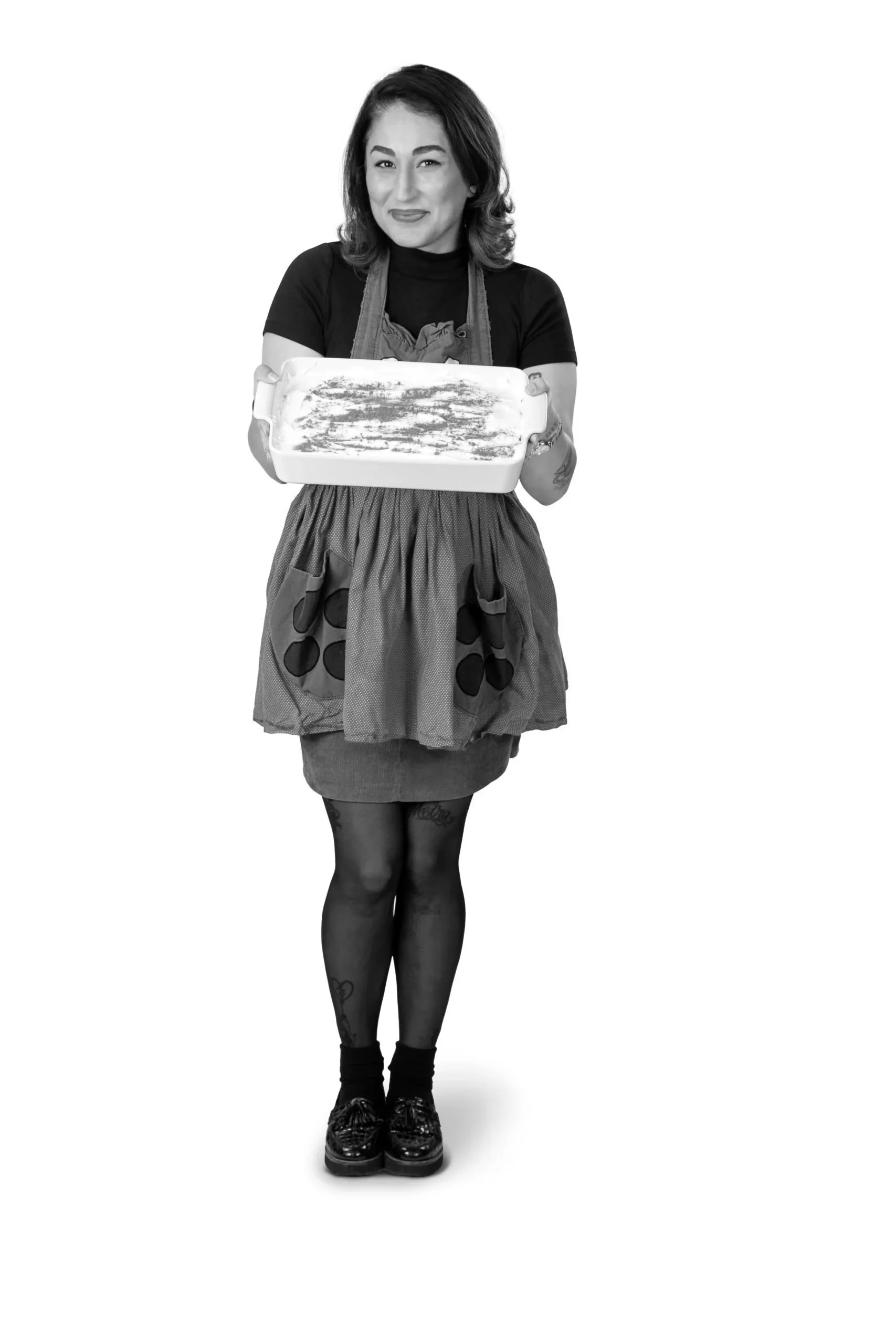 A woman in a black shirt and patterned apron holding an empty baking dish, standing against a white background.