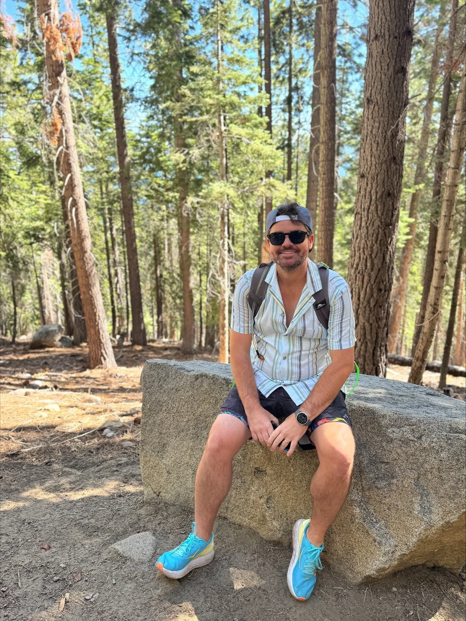 A man sitting on a large rock in a forest with tall trees and green foliage, wearing sunglasses, a baseball cap, a striped shirt, shorts, and running shoes.