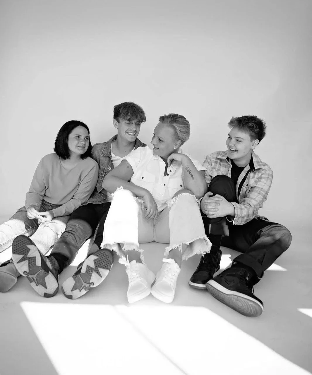 A group of four young people sitting on the floor against a plain background, smiling and interacting with each other, dressed casually.
