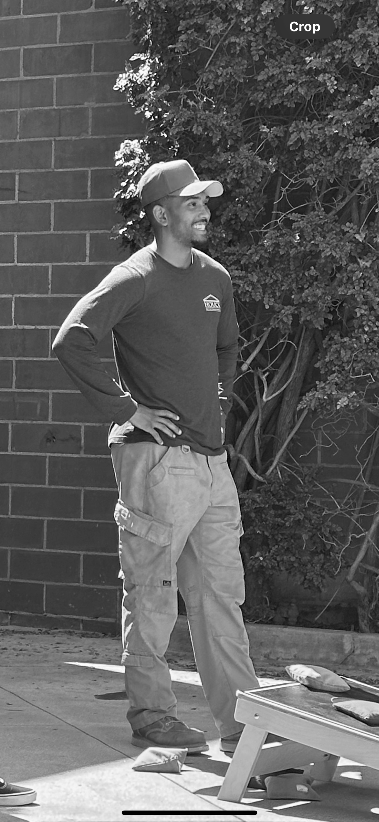 A man wearing a cap, long sleeve shirt with a logo, cargo pants, and sneakers standing outdoors on a sunny day, smiling with hands on his hips, next to a low table with some items on it, with a brick wall and vine-covered shrubbery in the background.
