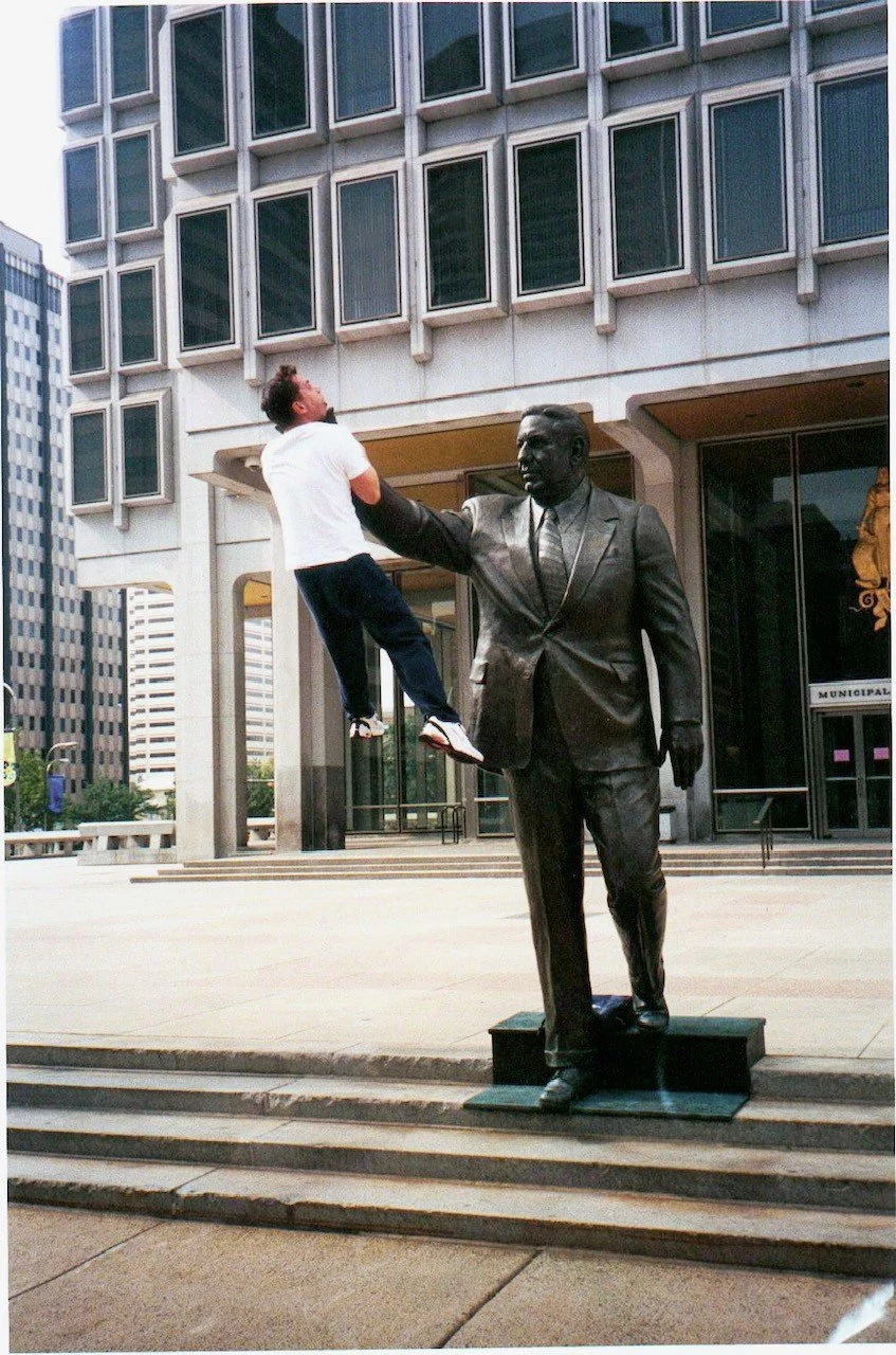 A person appears to be floating or flying while holding onto the outstretched hand of a statue of a man in a business suit, situated outside a building with a modern architectural facade.