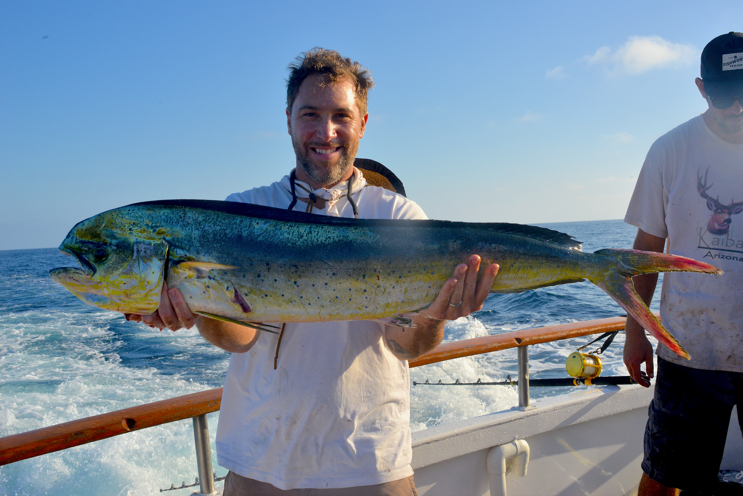 Man holding a large mahi-mahi fish on a boat at sea with another person in the background.