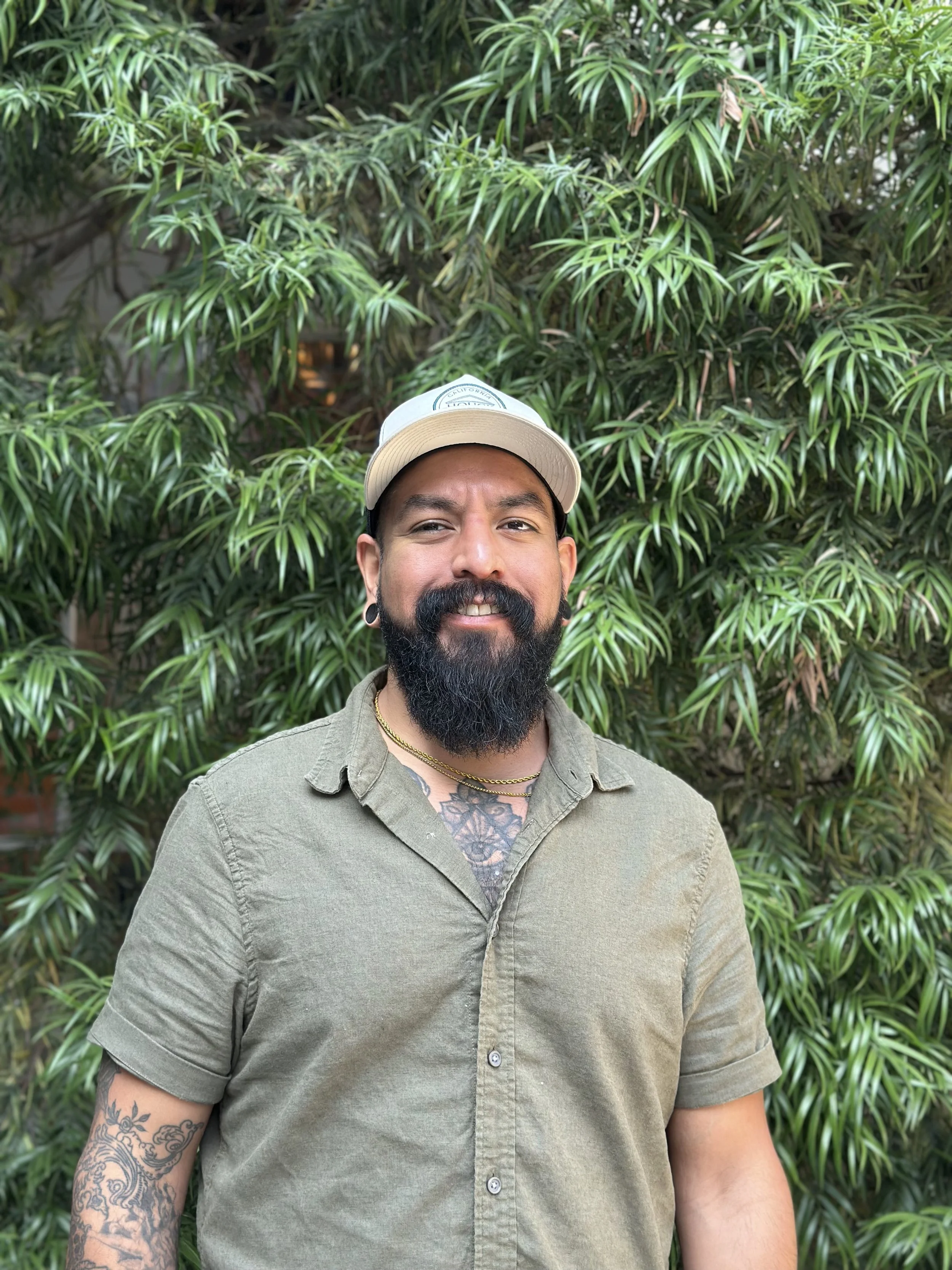 A man with a beard and tattoos, wearing a beige cap and a green button-up shirt, standing in front of lush green foliage.