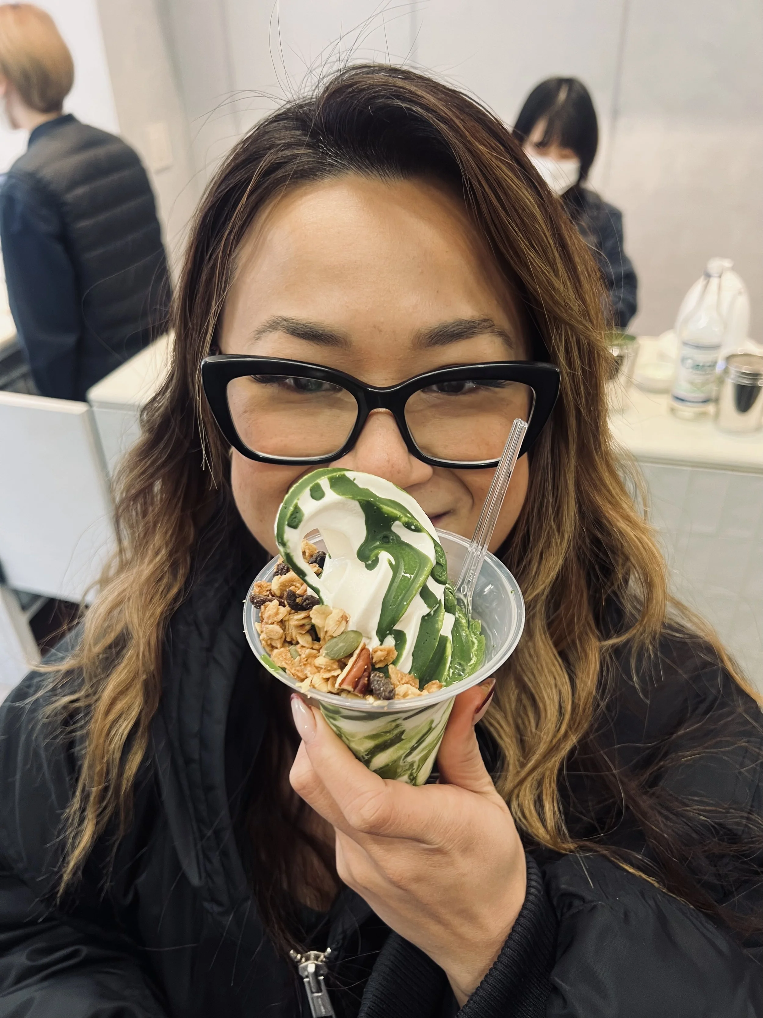A woman with glasses holding a cup of soft serve ice cream topped with granola and caramel drizzles in a cafe.